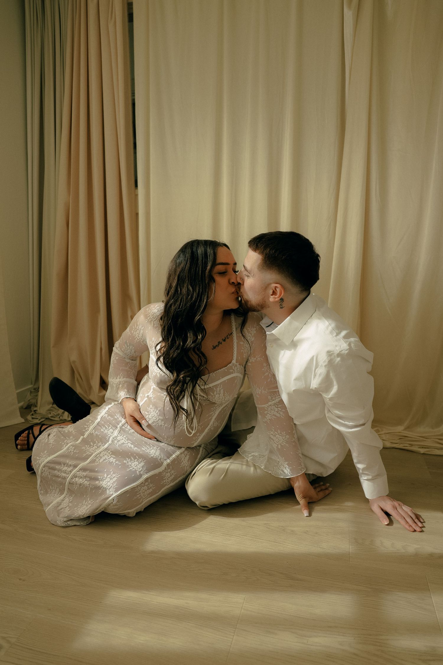 An intimate couple shares a tender moment on hardwood floor against cream curtains.