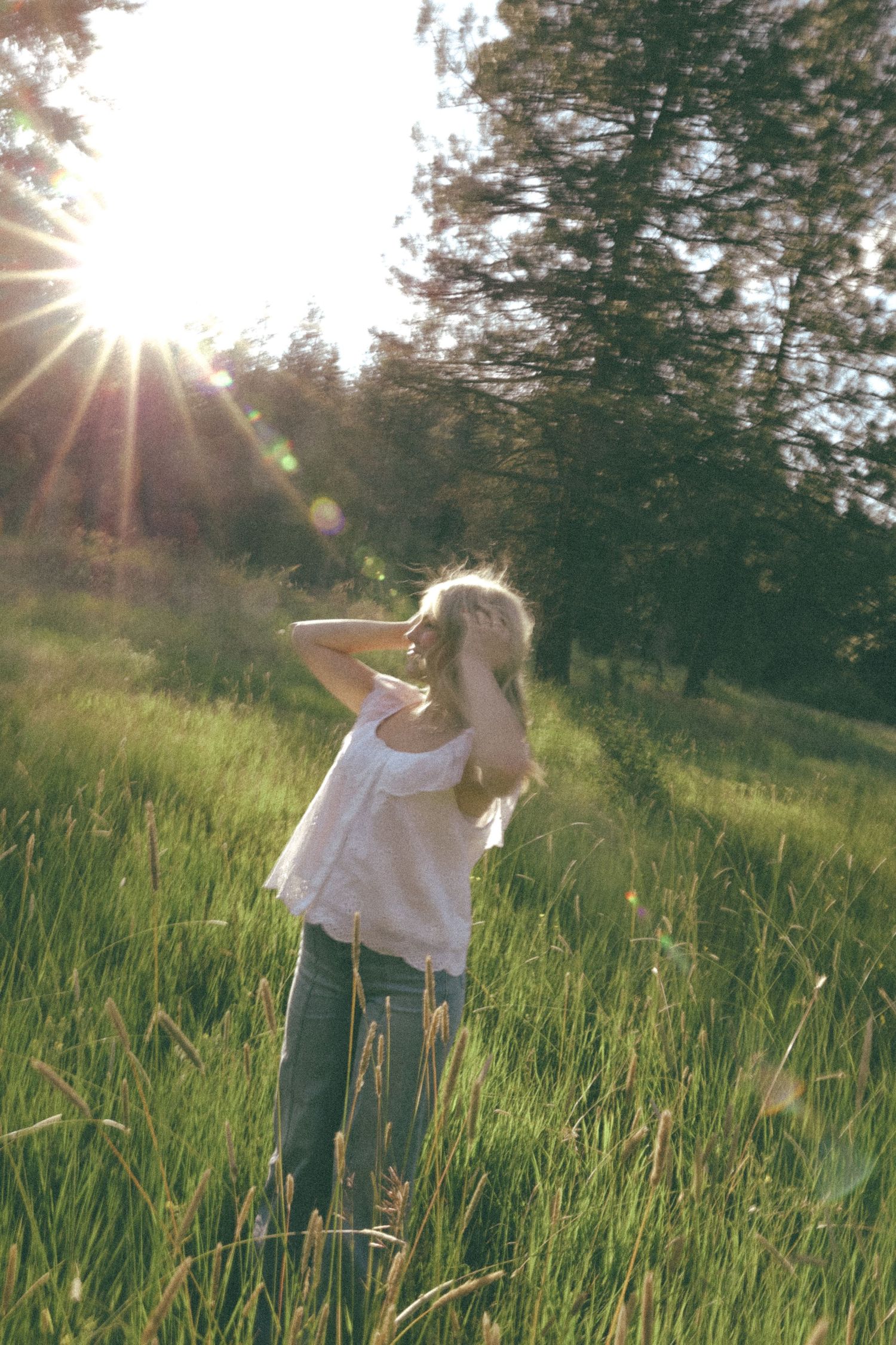 Person dancing gracefully in a sunlit meadow with tall green grass and evergreen trees in the background.