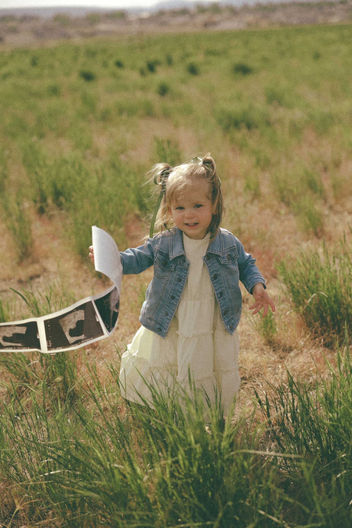 A young girl in denim plays with a white toy in tall grass.