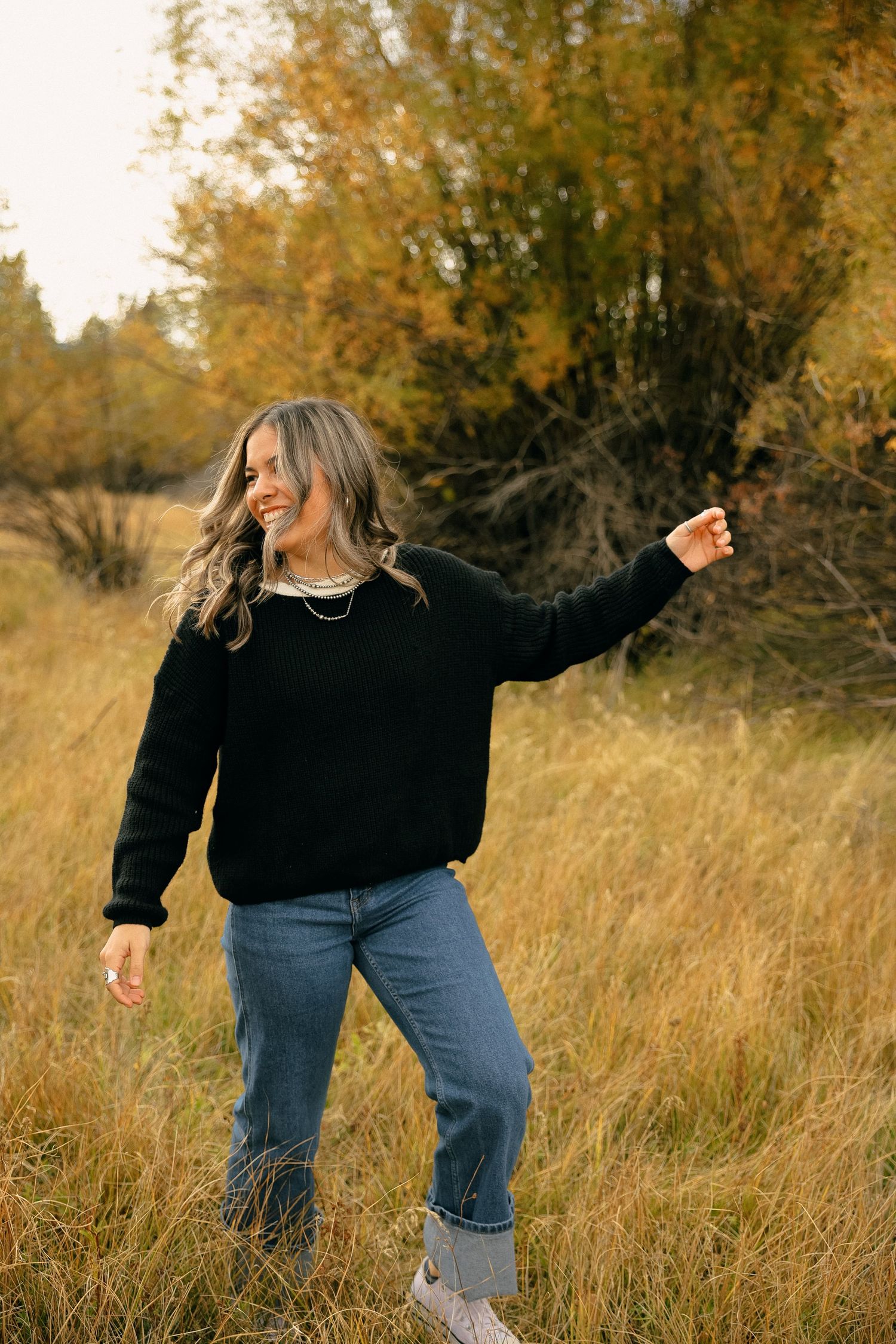 Person in black sweater and jeans dances joyfully in tall autumn grass.