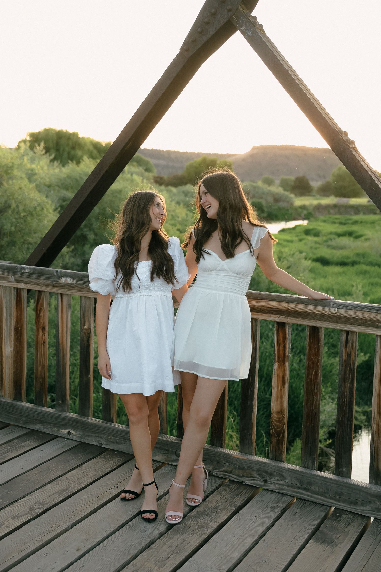 Two friends in white dresses share a moment on wooden bridge at sunset.