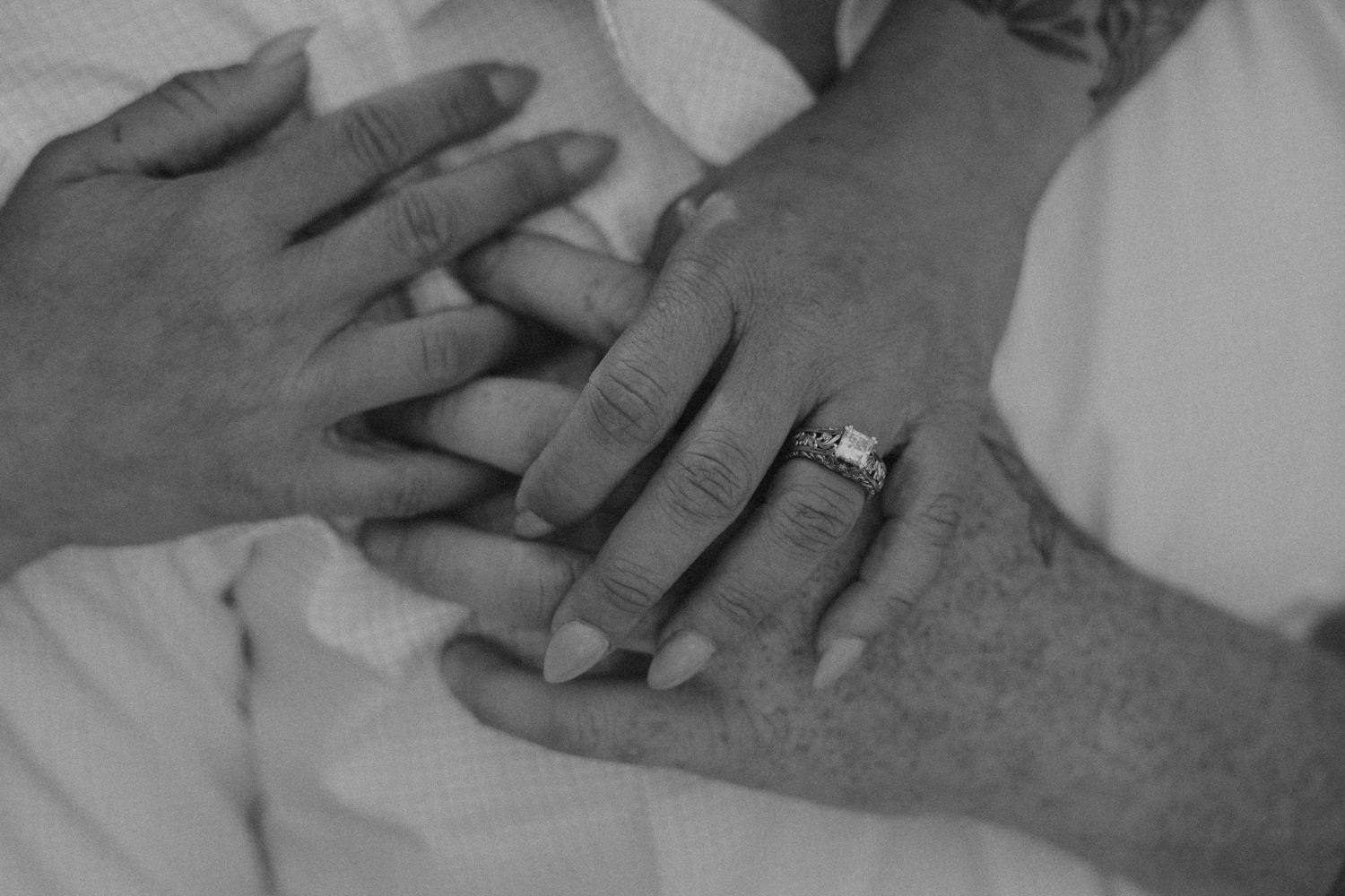A black and white close-up photograph shows hands with a diamond ring resting on white fabric.