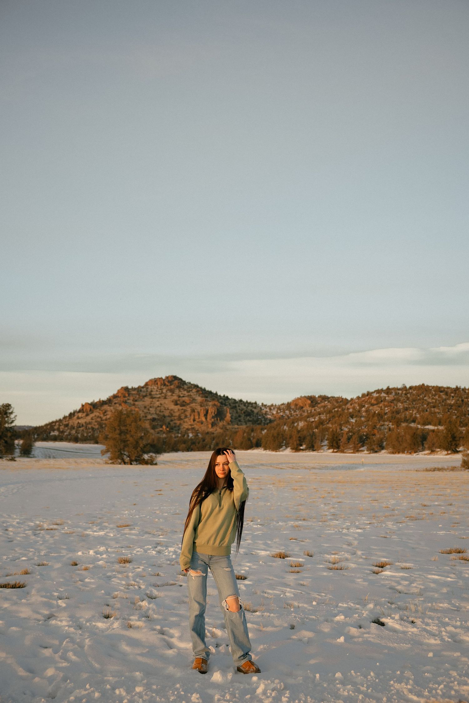 Person stands in snowy landscape with mountainous backdrop at sunset, wearing casual winter attire.