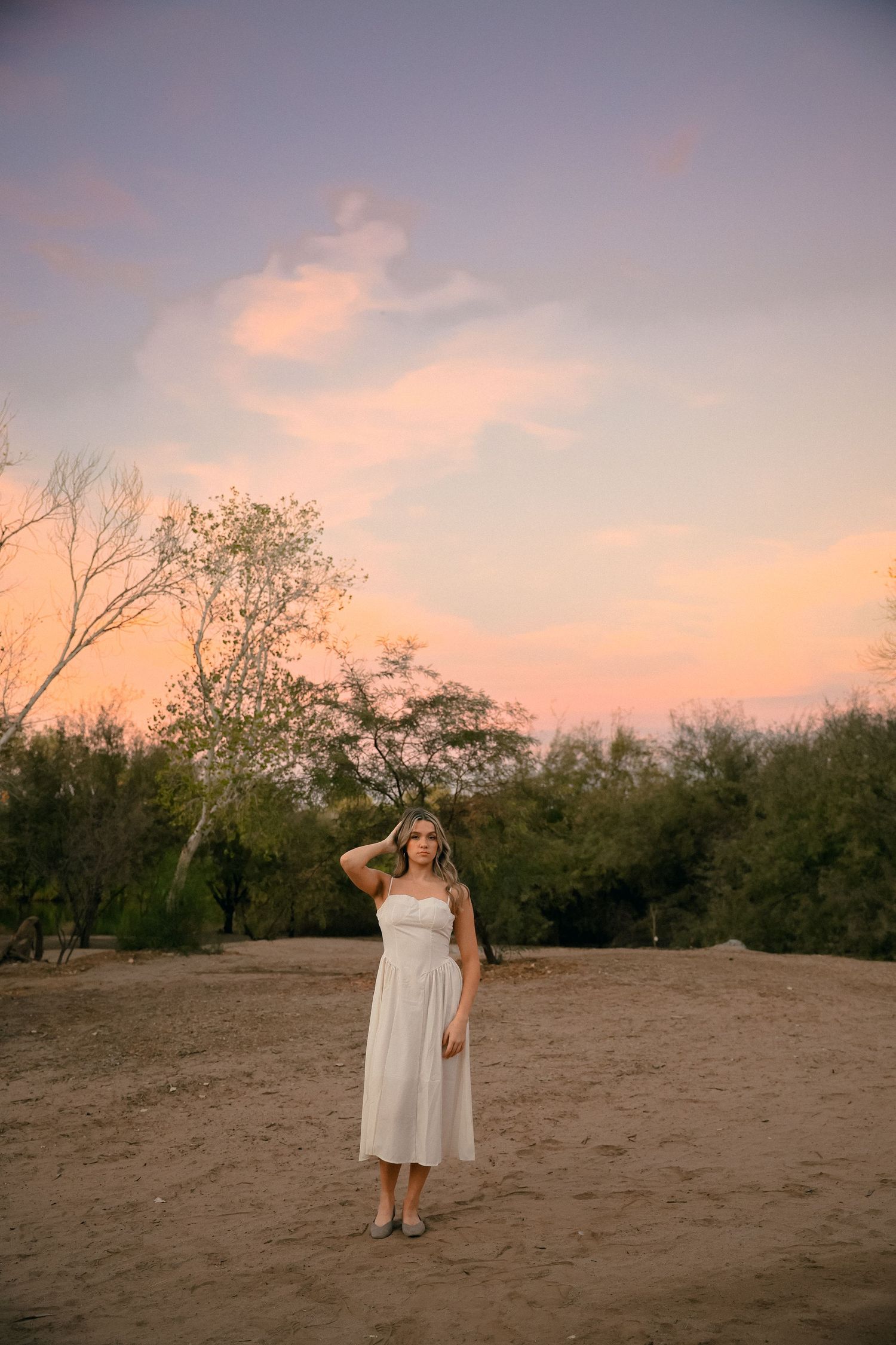 A woman in a white dress poses in a desert landscape during a pink sunset.