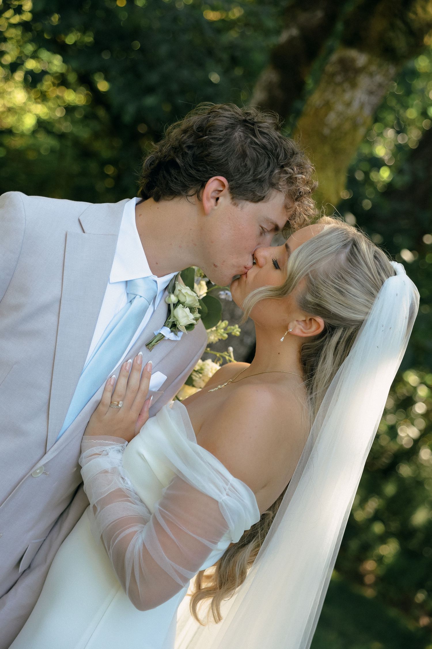 A romantic wedding kiss captured in a garden setting with light filtering through trees.