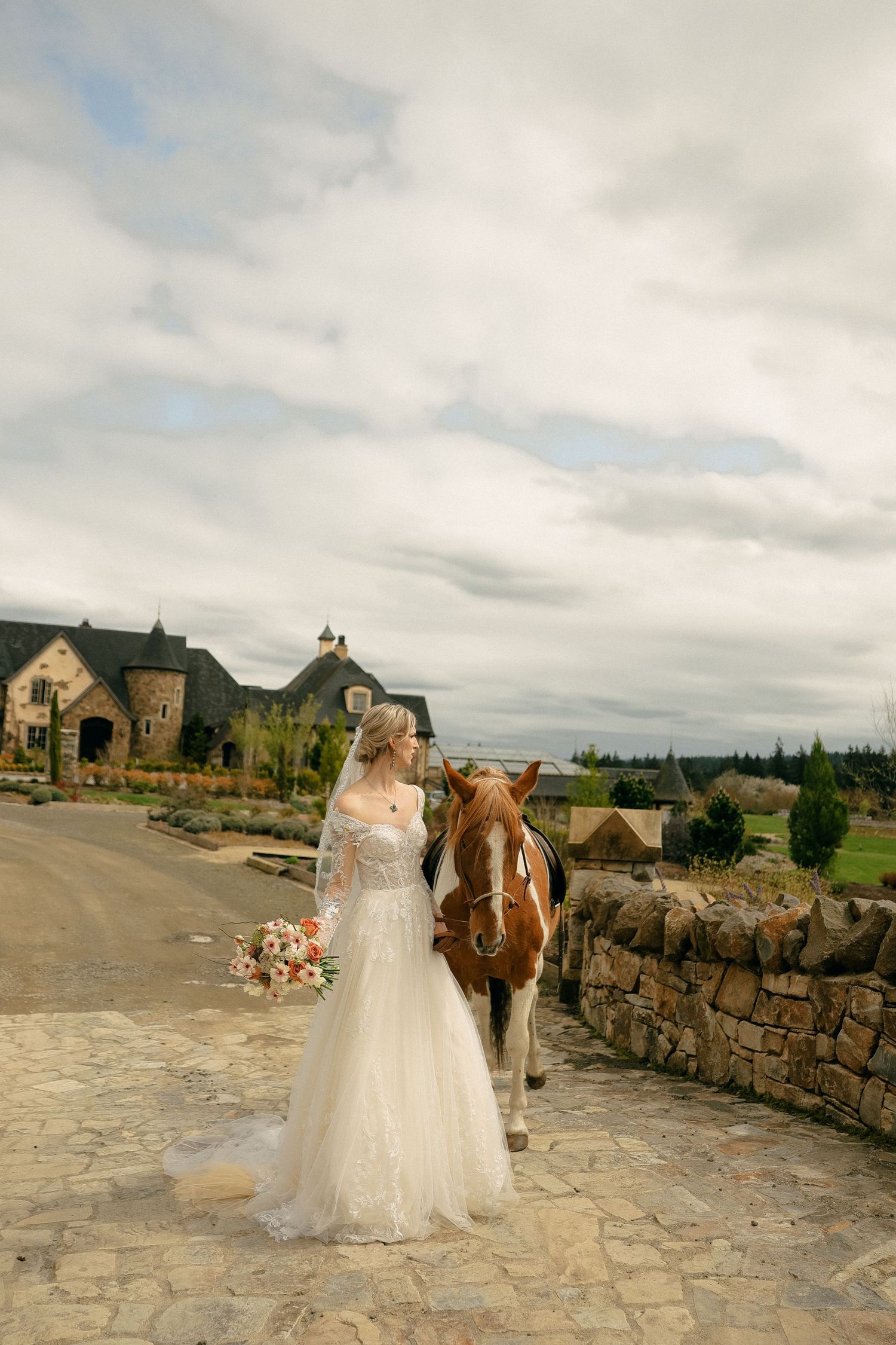 Bride in flowing lace gown poses with horse outside rustic stone mansion.