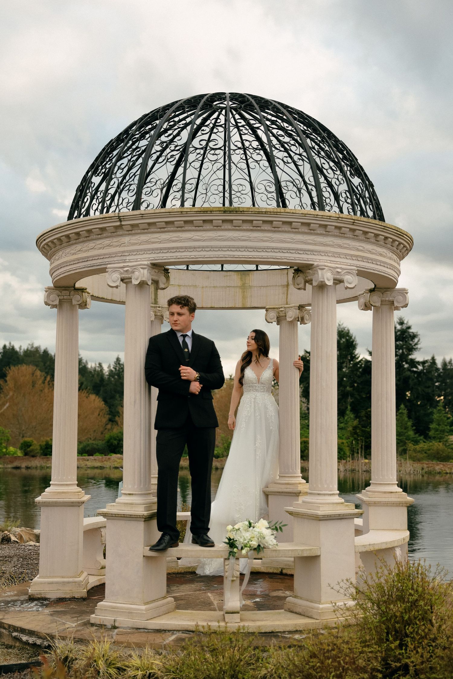Elegant couple stands in white domed gazebo overlooking waterfront during wedding ceremony.