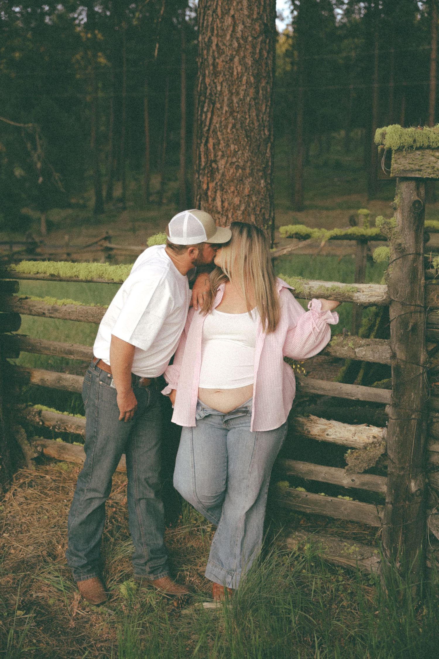 Two people in casual attire lean against a rustic wooden fence surrounded by tall pine trees in a forest setting.