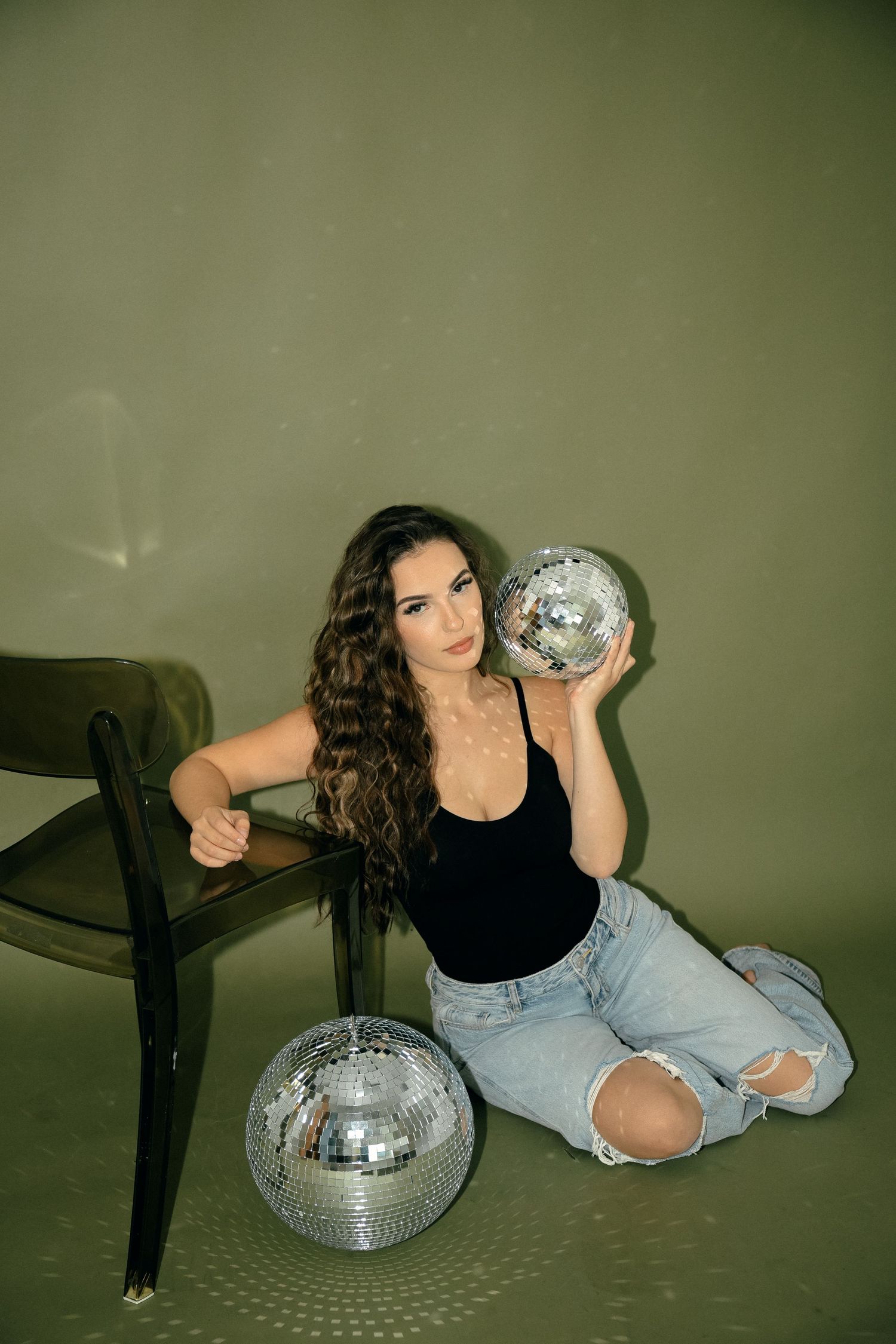 Person poses with disco balls on floor wearing casual attire against green wall.