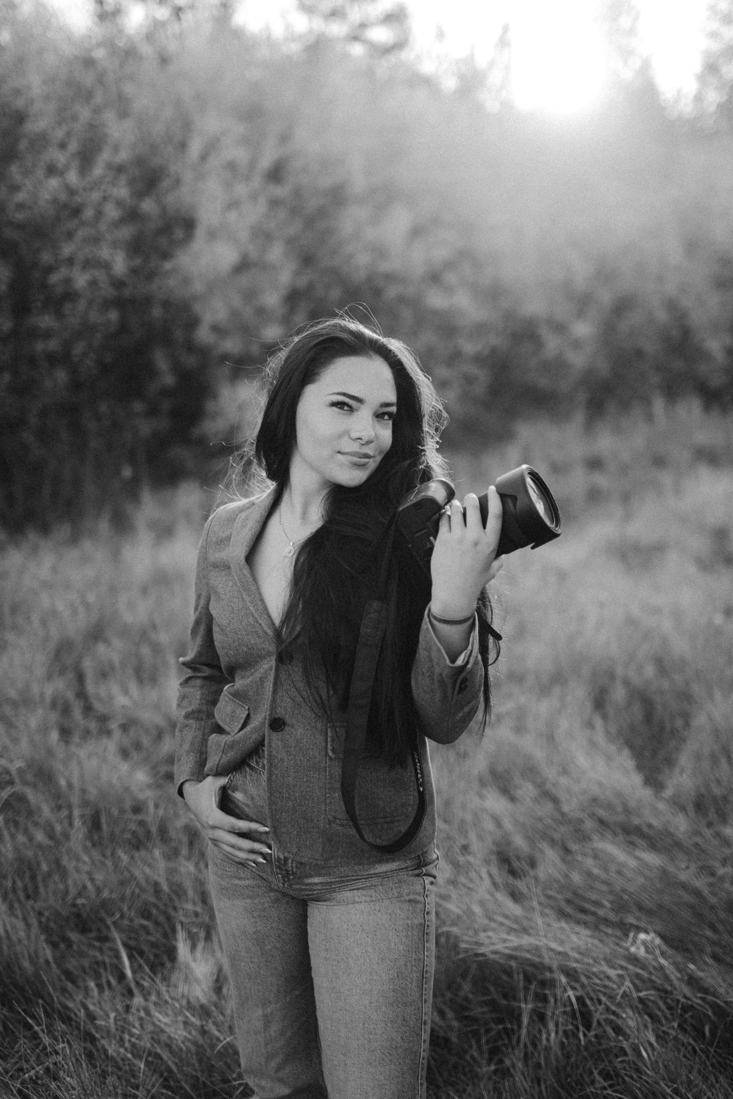 Professional photographer in casual attire holding camera in sunlit meadow in black and white.