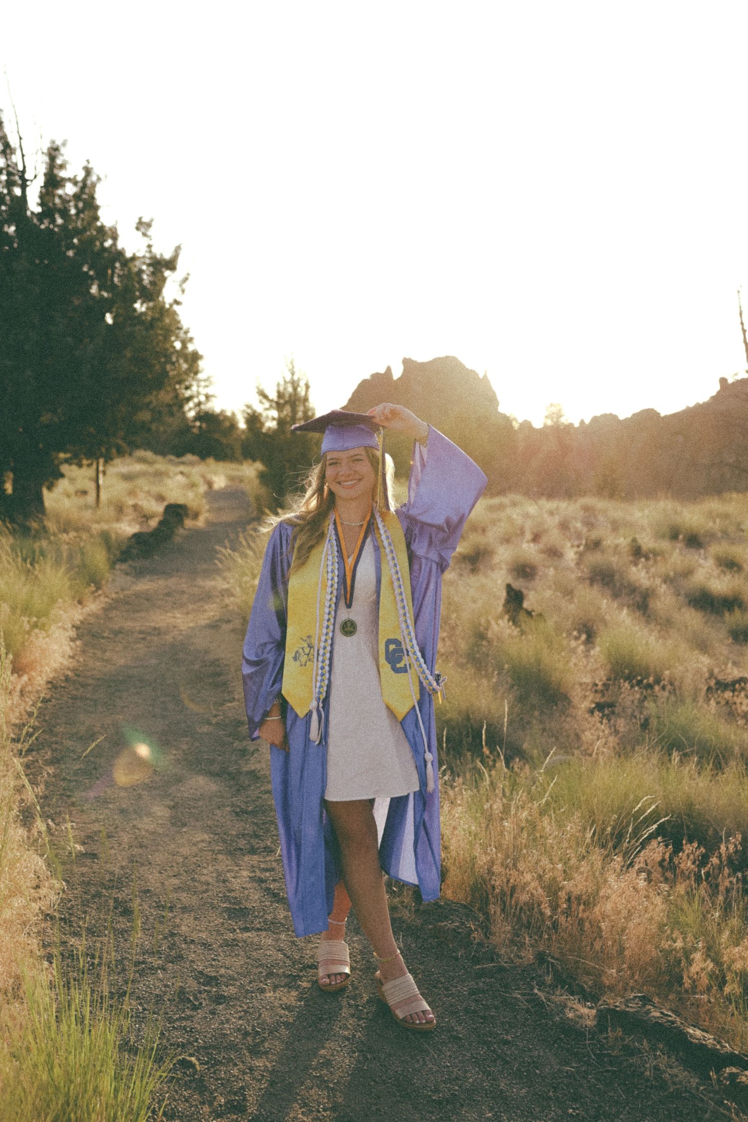 A graduate in purple and gold regalia walks along a sunlit desert trail during golden hour for graduation photos.