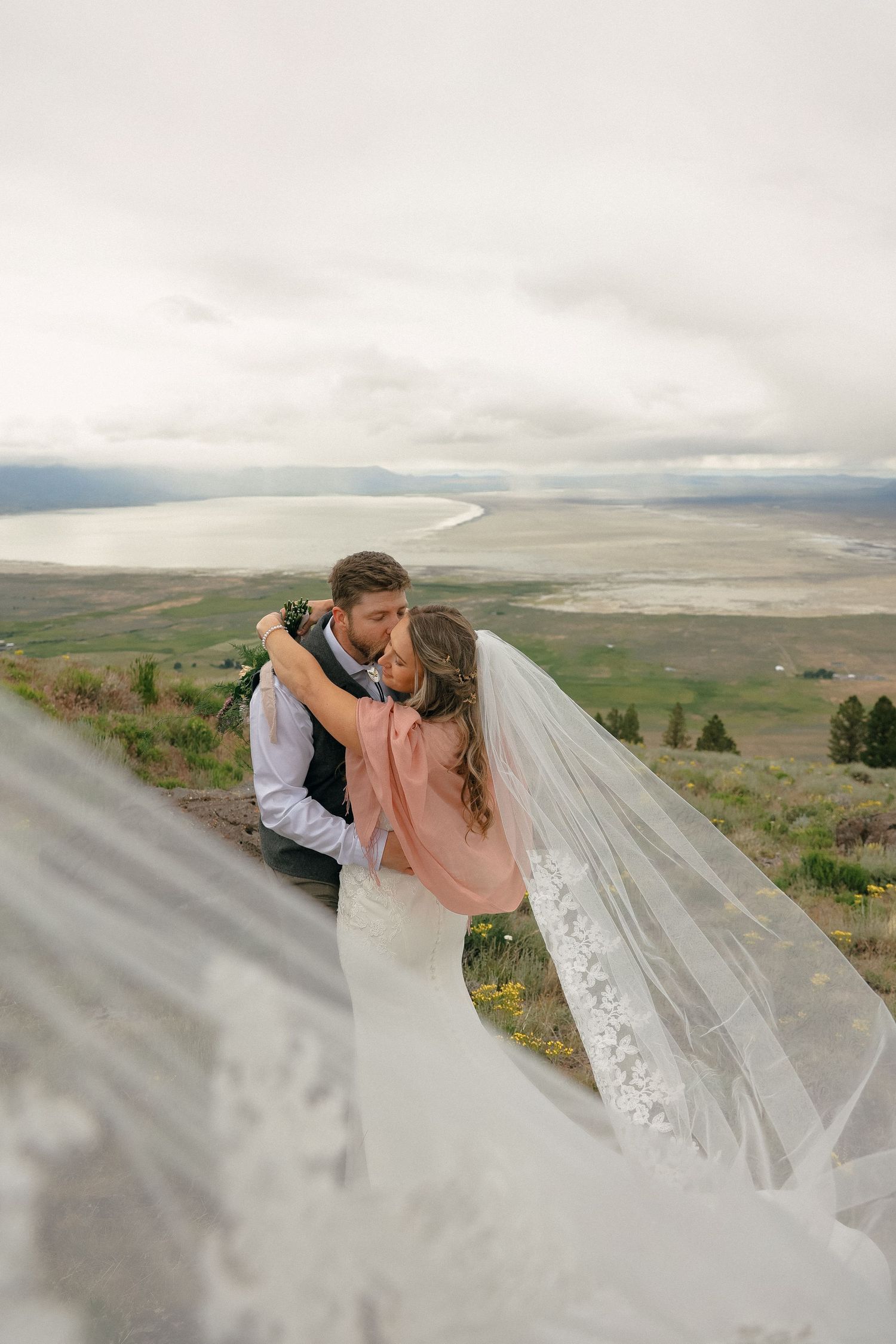 A couple in wedding attire embrace under a flowing veil against a scenic mountain landscape backdrop.