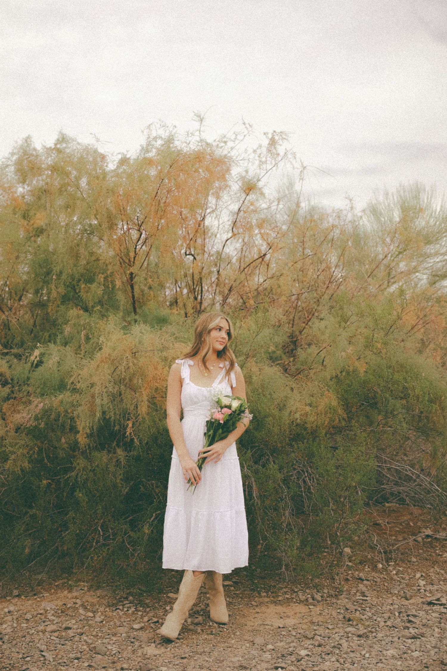 A woman in a white sundress and boots stands holding flowers against an autumn backdrop.