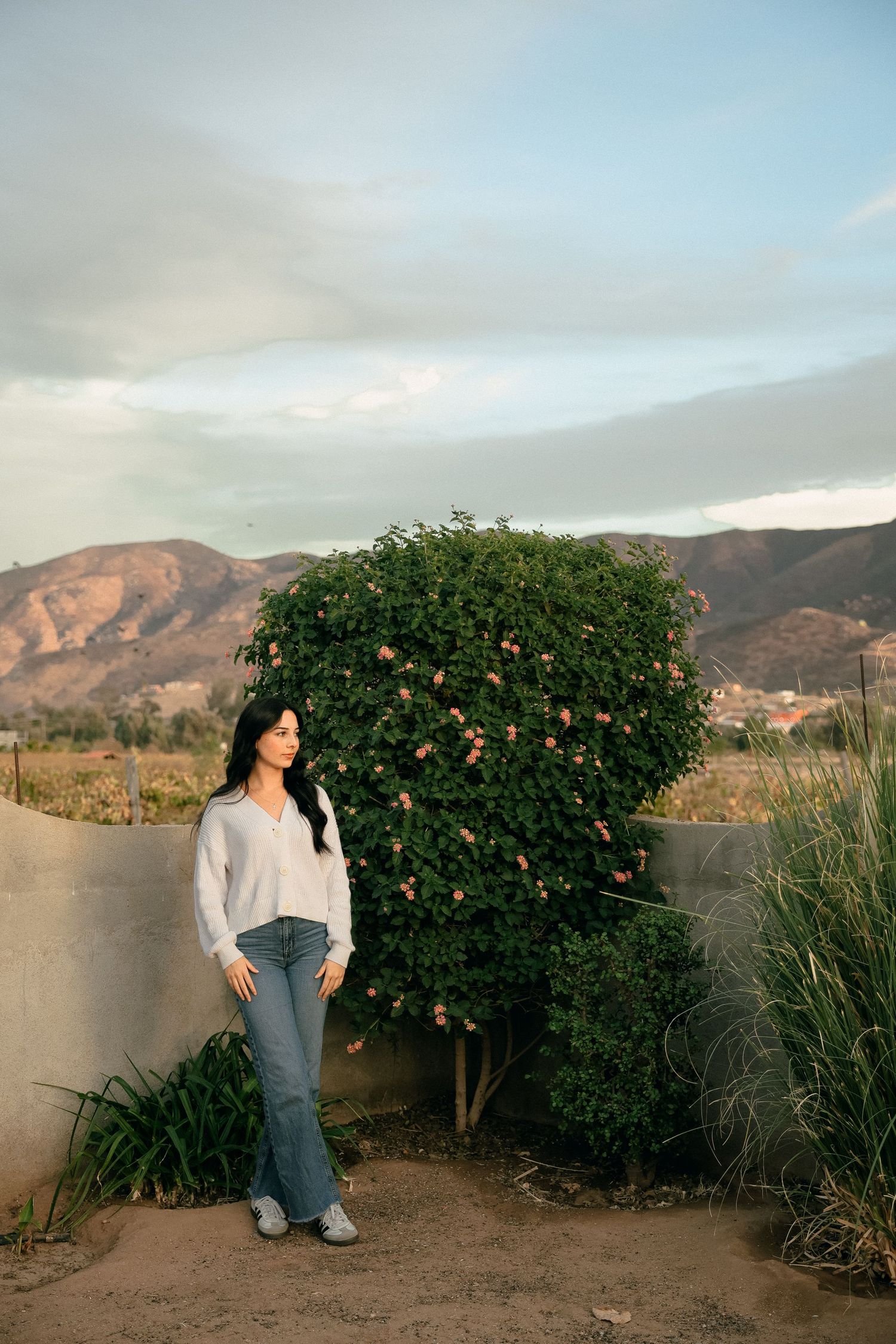 Woman in casual attire poses beside large flowering bush with mountains in background.