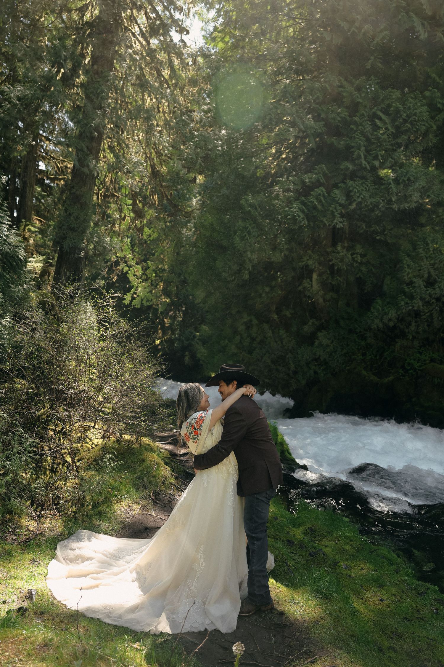 Wedding couple embracing beside rushing waterfall in mossy forest setting.