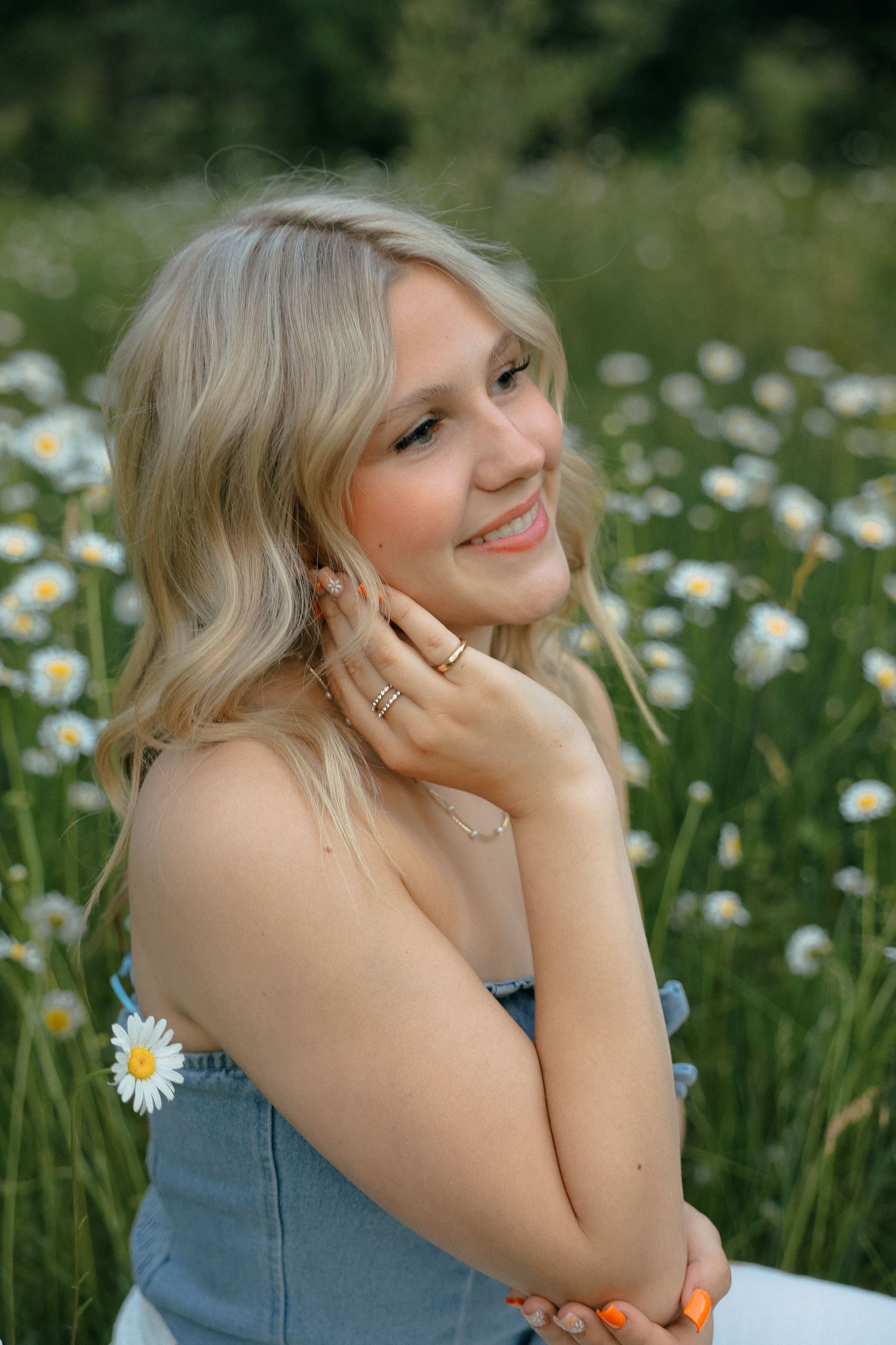 Blonde woman in denim dress poses in a sunlit meadow of white daisies with a gentle smile and hand near face.