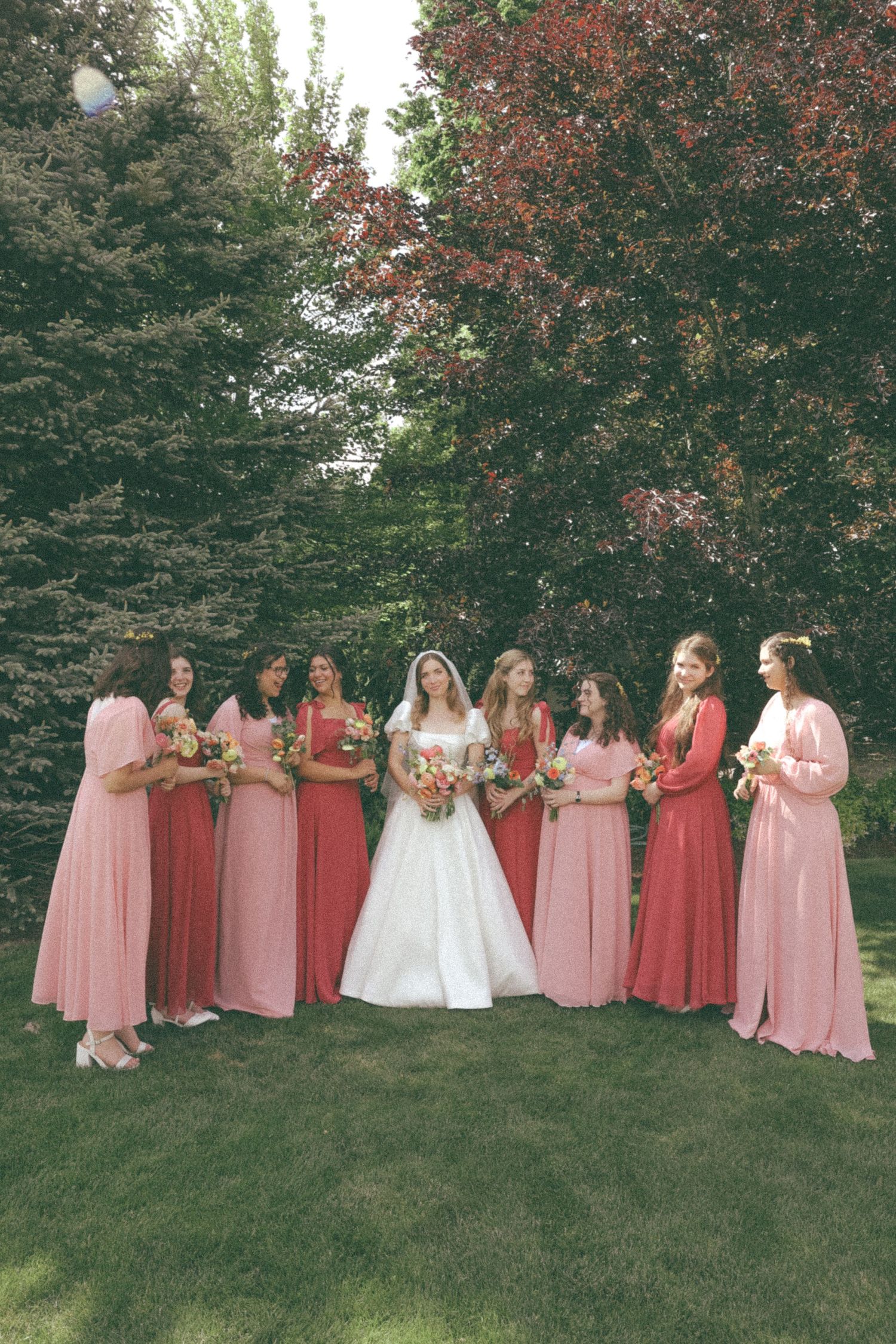 Bridesmaids in pink dresses pose with bride on grassy hill.
