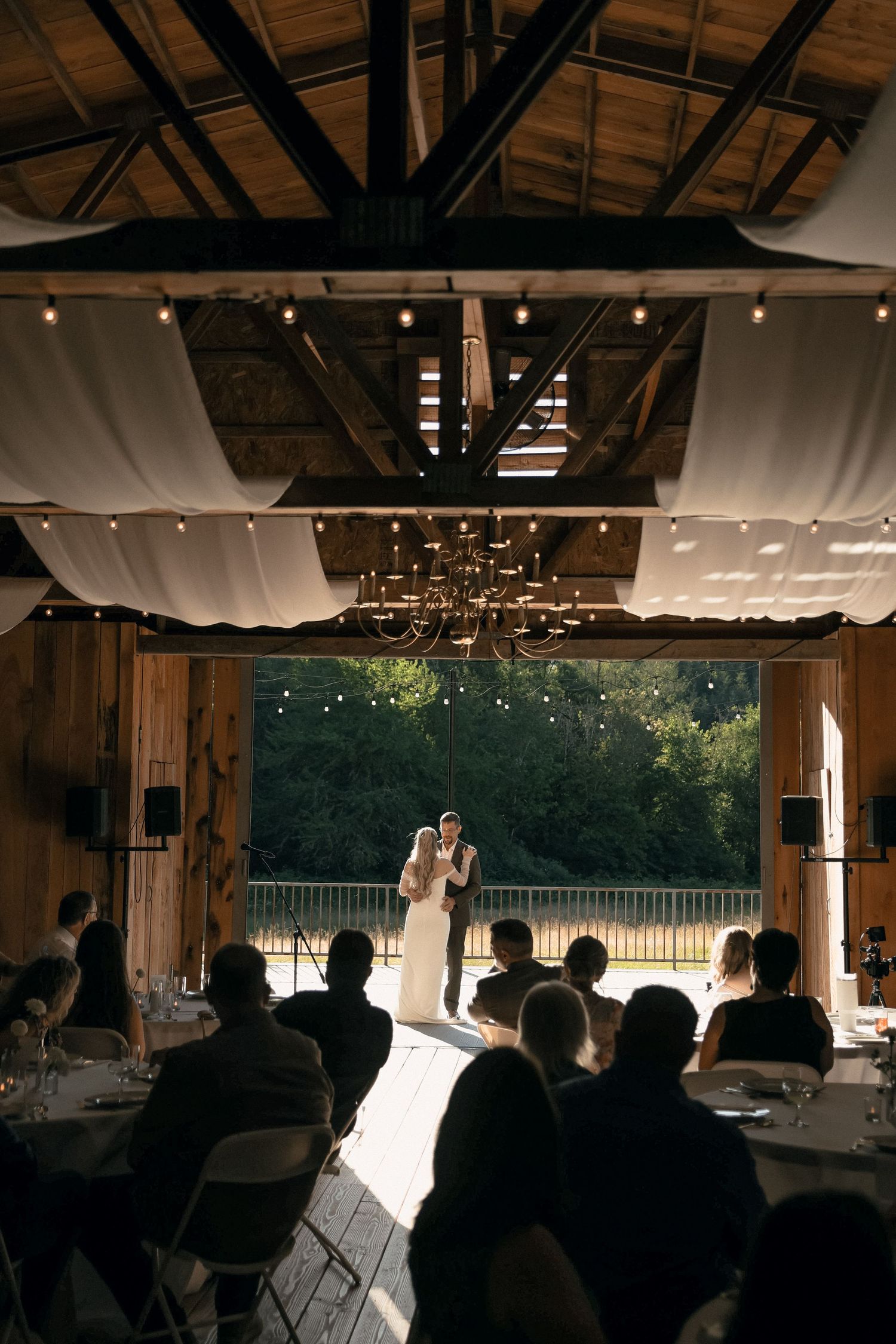 Indoor rustic wedding venue with draped fabric ceiling, string lights, and silhouetted guests watching a couple on stage.