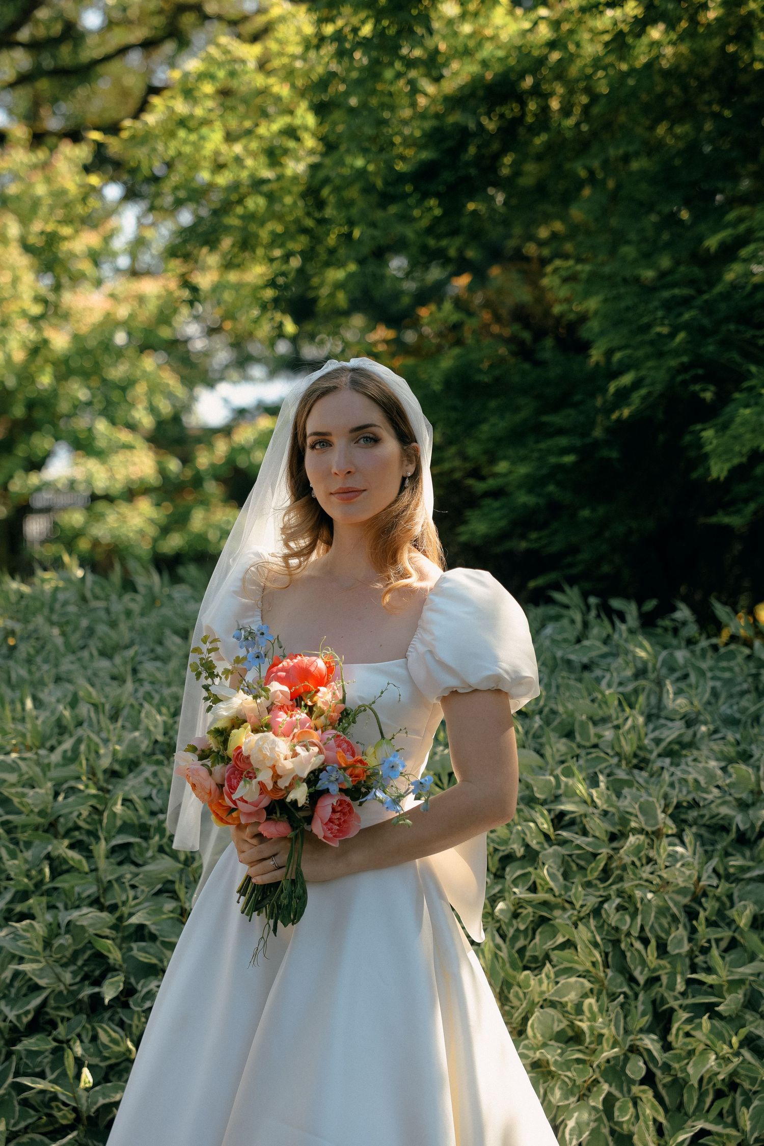 Bride in elegant white gown holding coral and peach bouquet in garden setting.