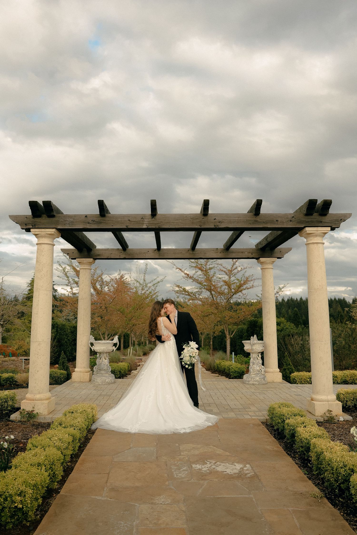 Wedding couple poses under a classical stone pergola in a manicured garden setting.