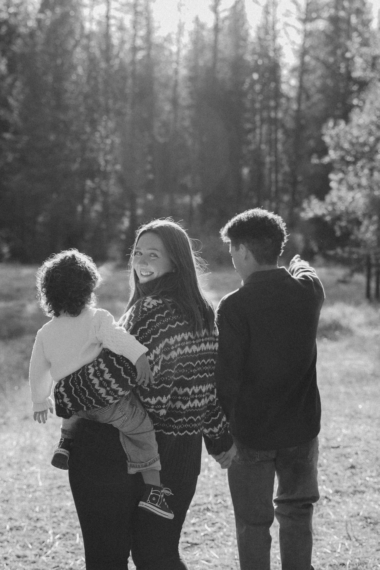 A family of three stands together in backlit forest setting wearing matching knit sweaters.