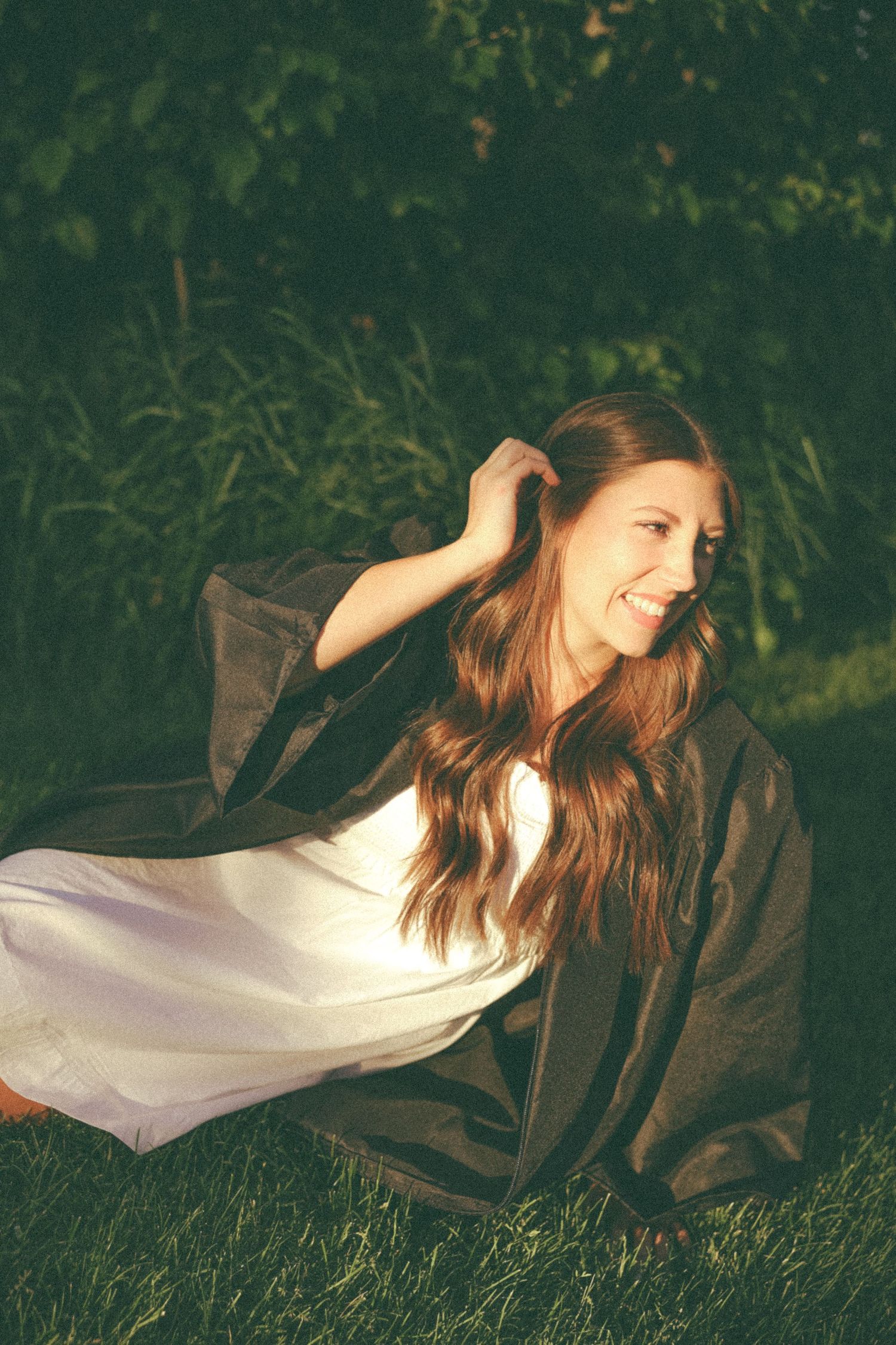 Person in white dress and dark jacket reclines on grass in evening light.
