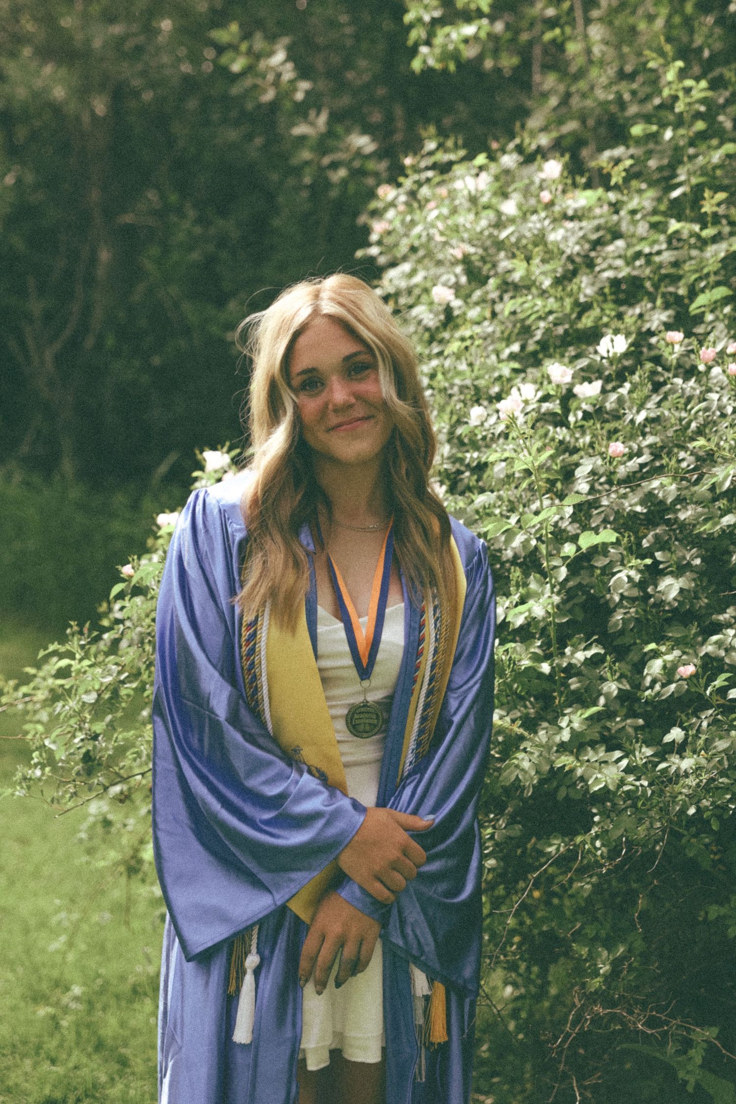 Graduate in blue cap and gown smiling outdoors with flowering bushes in background.