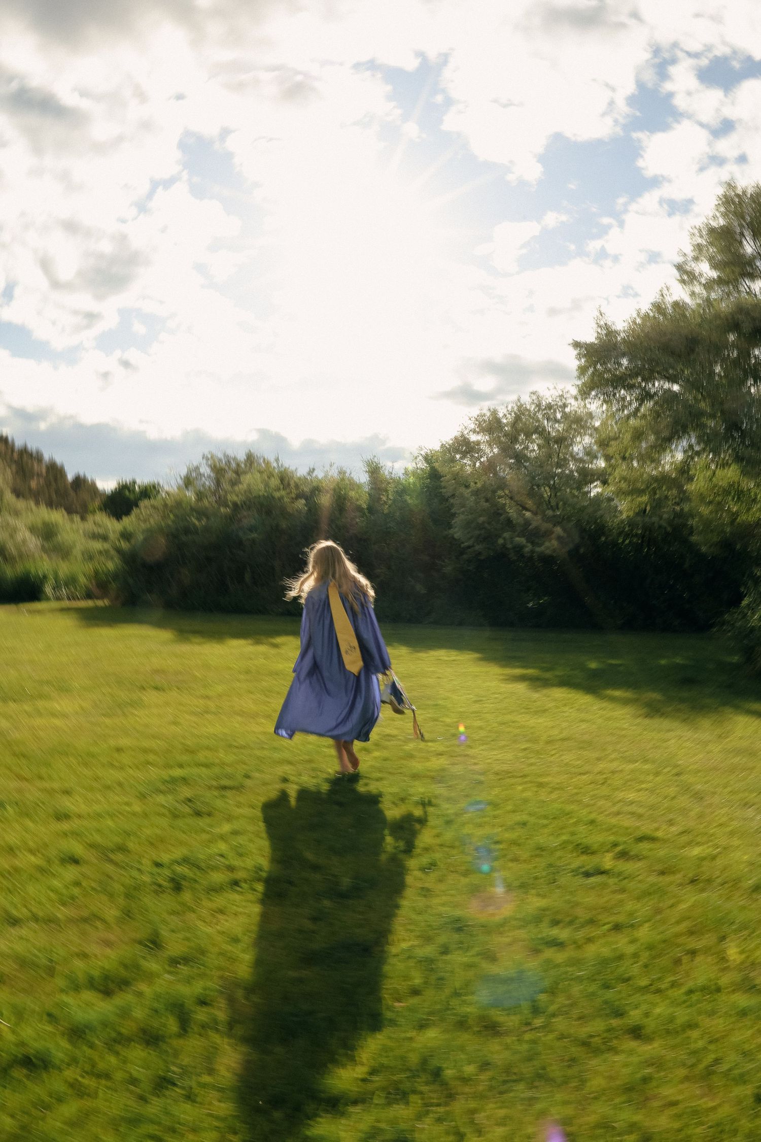 A figure in a flowing blue dress jumps in a sunlit meadow surrounded by trees on a cloudy summer day.