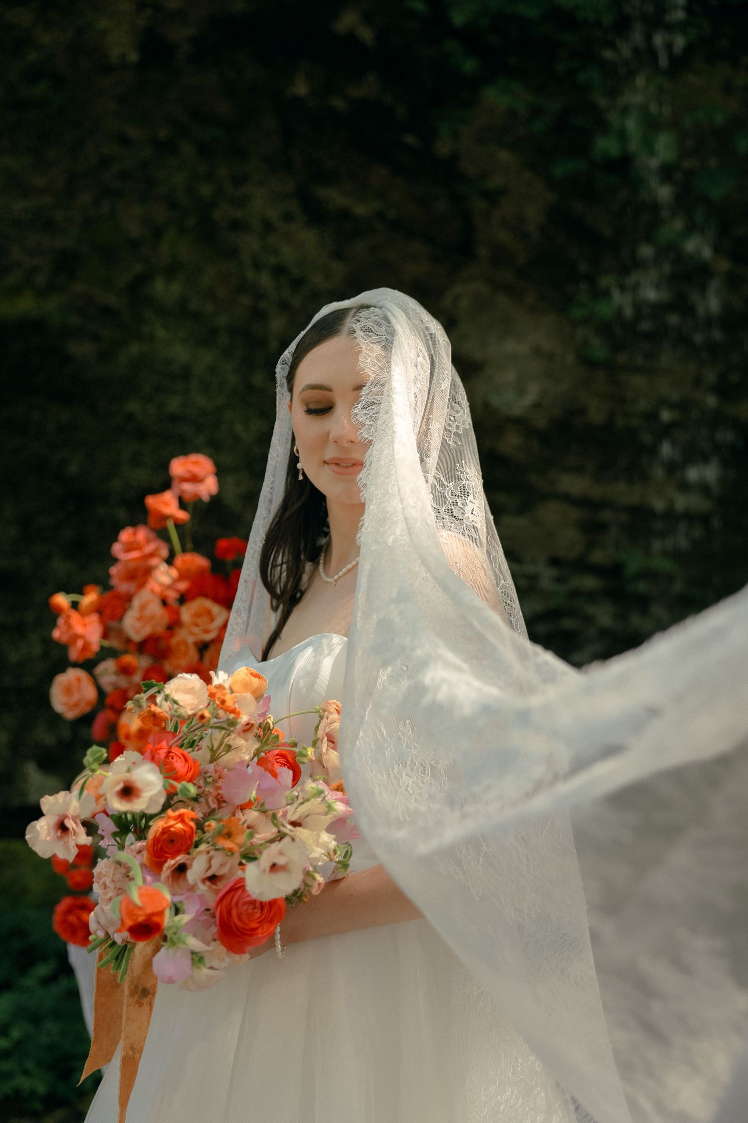 Bride in lace veil holds vibrant coral and peach floral bouquet against garden backdrop.
