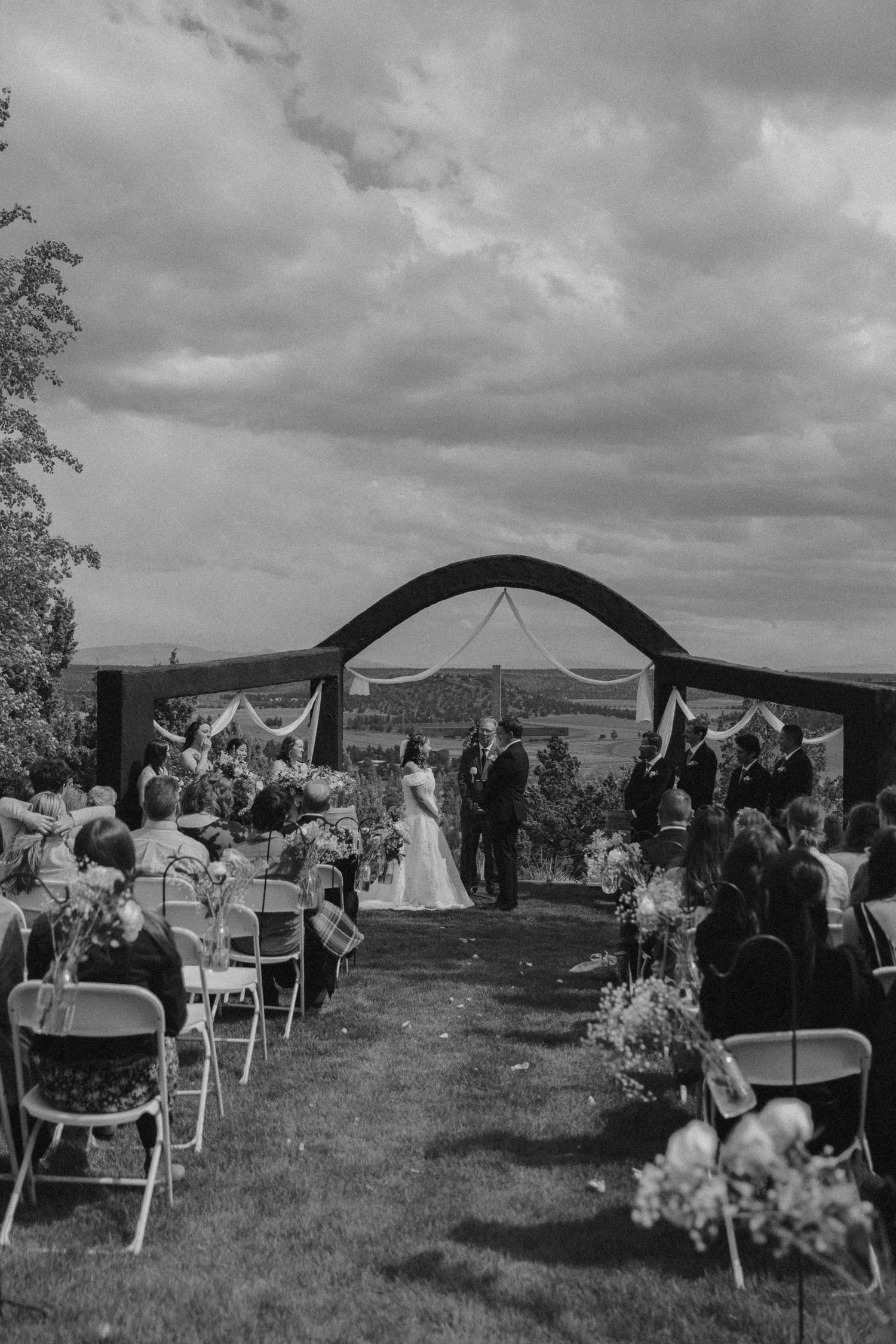 Black and white photo of outdoor wedding ceremony setup under cloudy sky.
