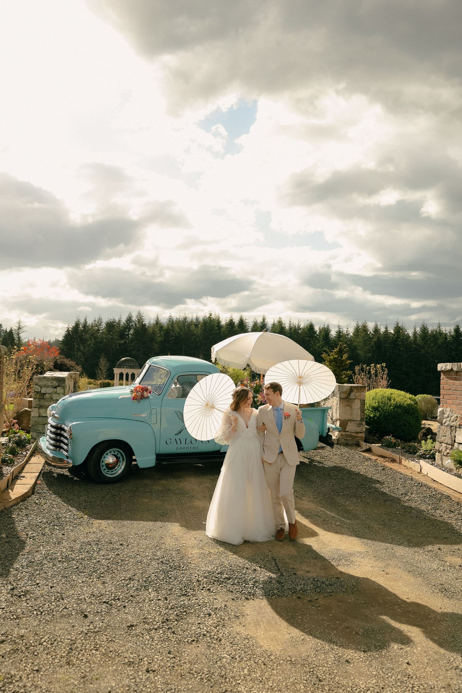 A vintage blue pickup truck serves as backdrop for a couple with white parasols on their wedding day.