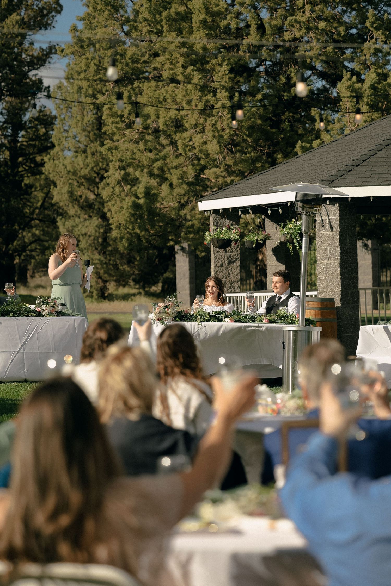 Outdoor wedding reception under a white tent with guests seated at tables during a meal.
