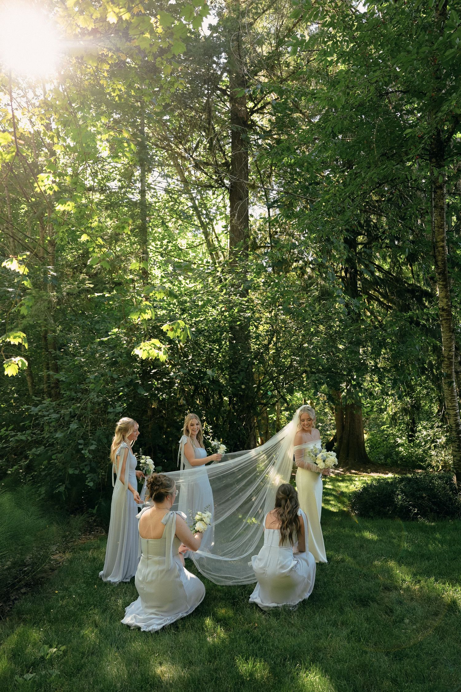 A magical woodland scene shows bridesmaids helping with a flowing wedding veil as sunlight filters through the trees.