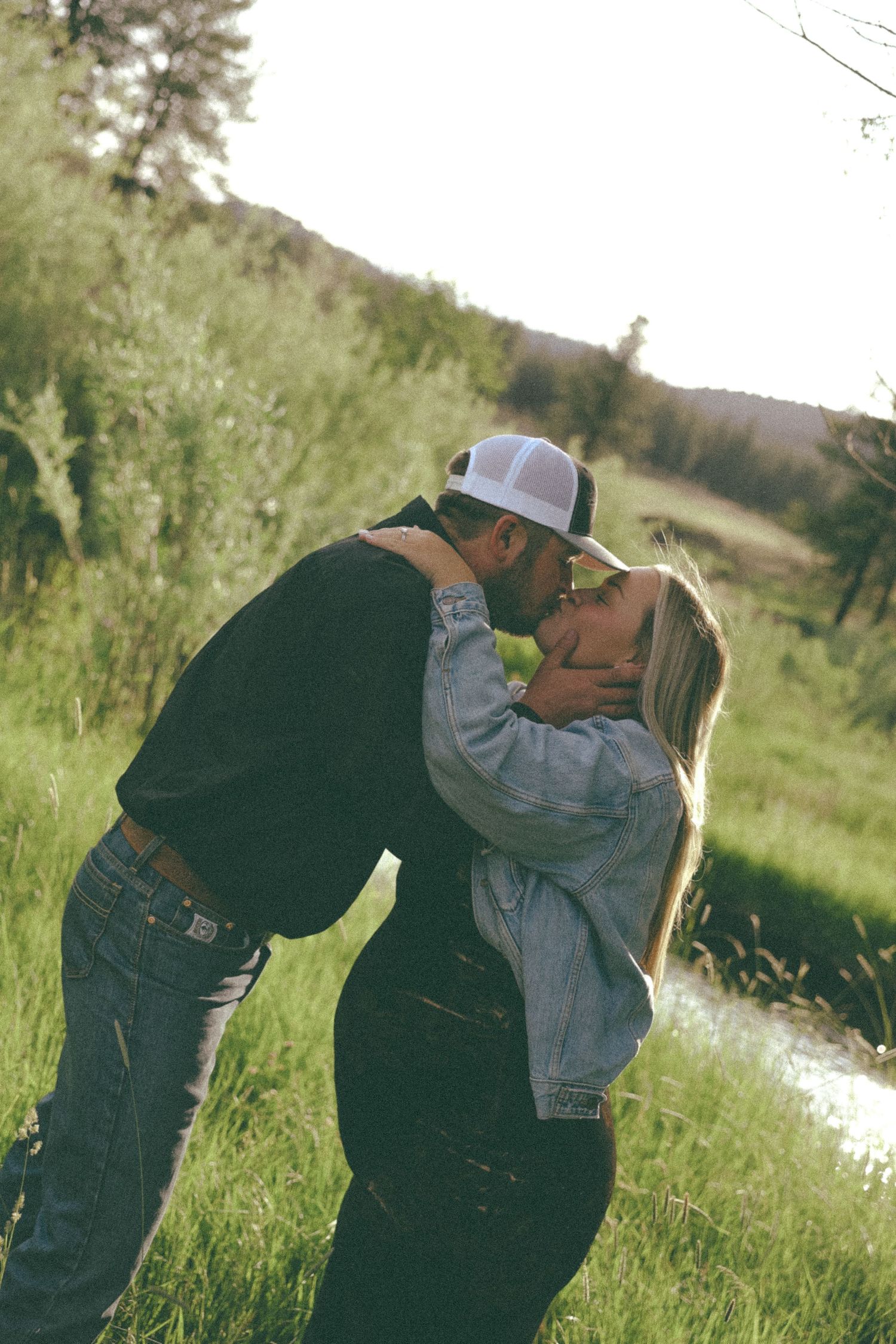 A romantic outdoor portrait shows a couple kissing in a sunlit meadow with rolling hills in the background.