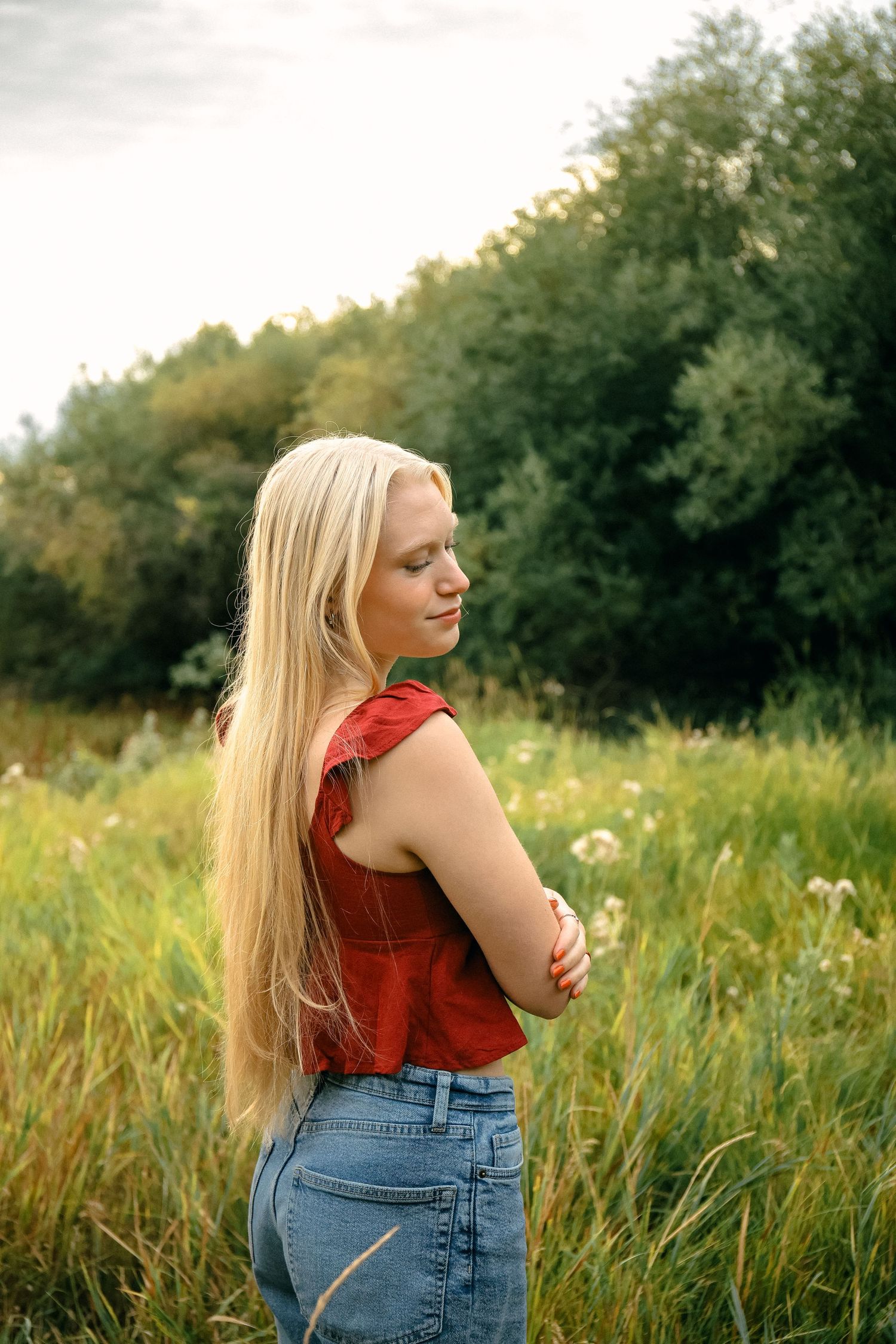A woman in a red sleeveless top and jeans stands in a sunlit meadow with wildflowers and trees in the background.