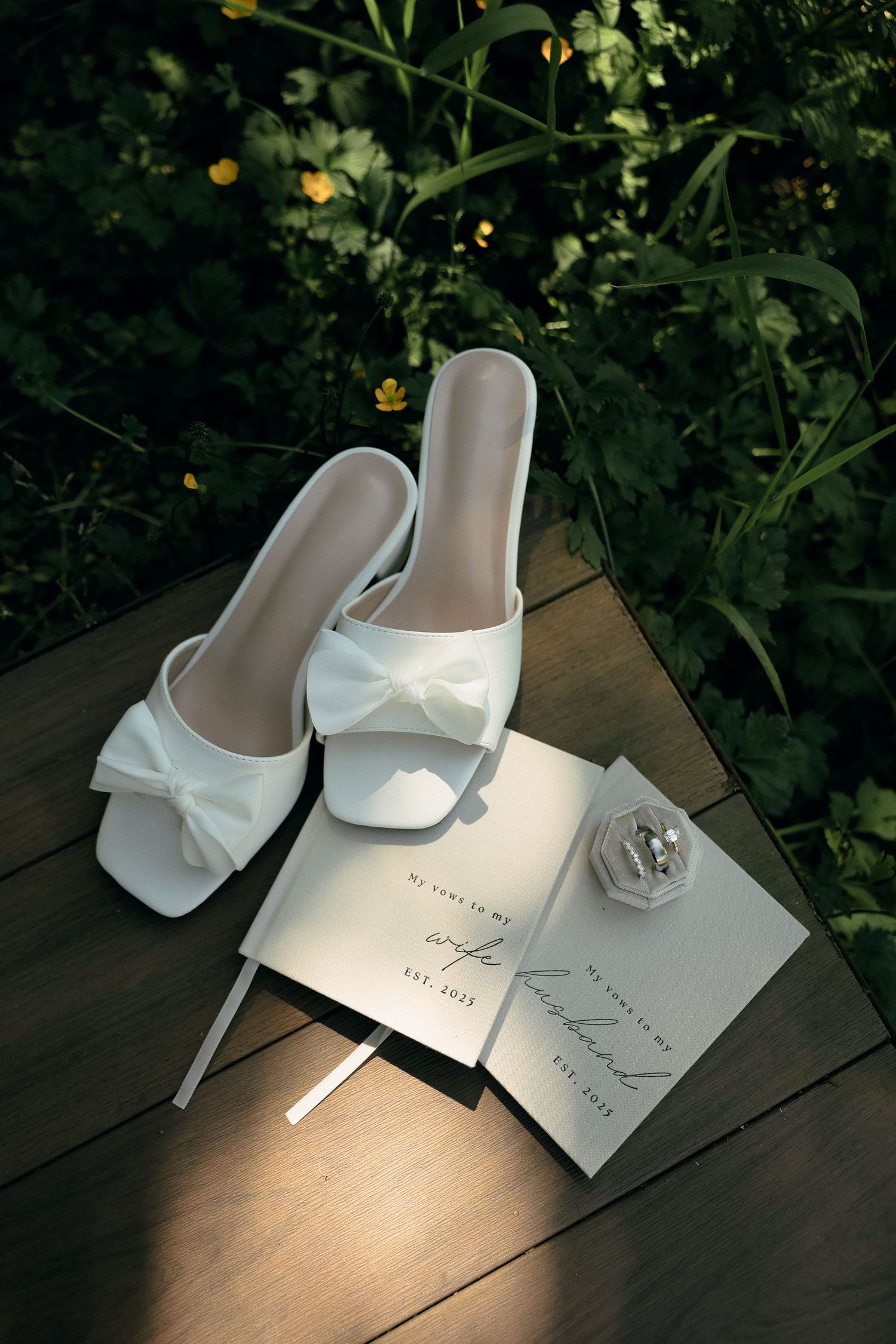 White bridal slide sandals with bows sit beside wedding invitation cards on a wooden surface in dappled sunlight.