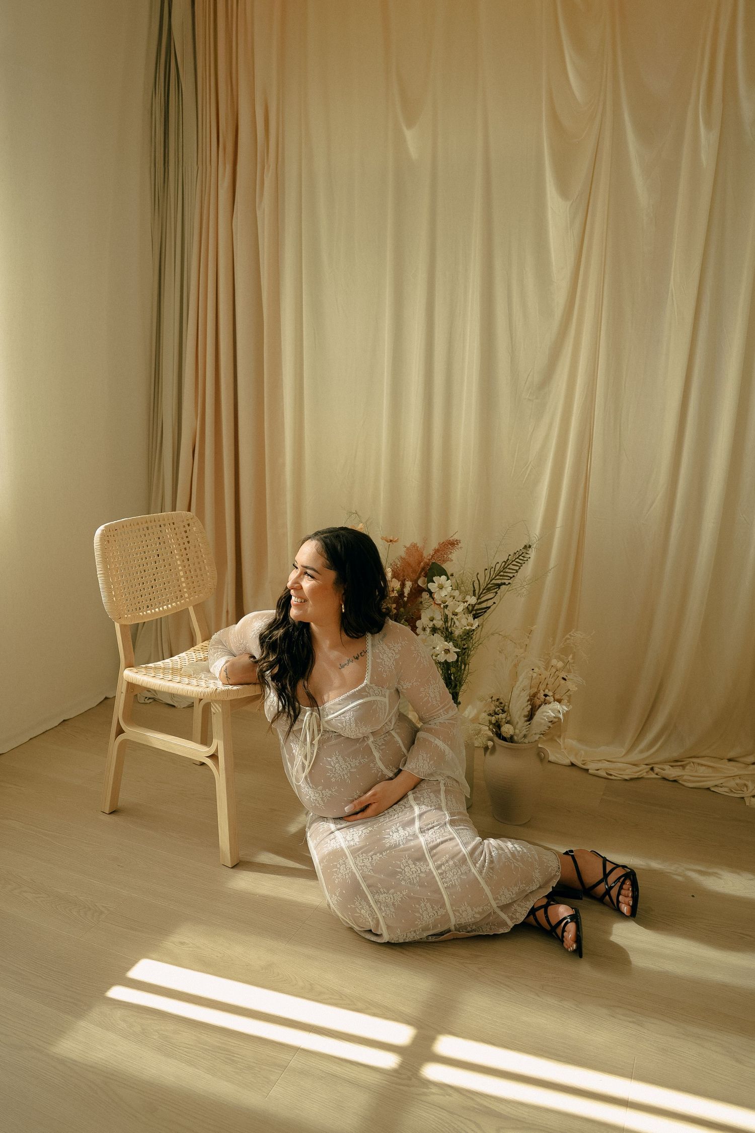 A woman in a white lace dress sits on the floor beside a rattan chair in a minimalist studio setting.