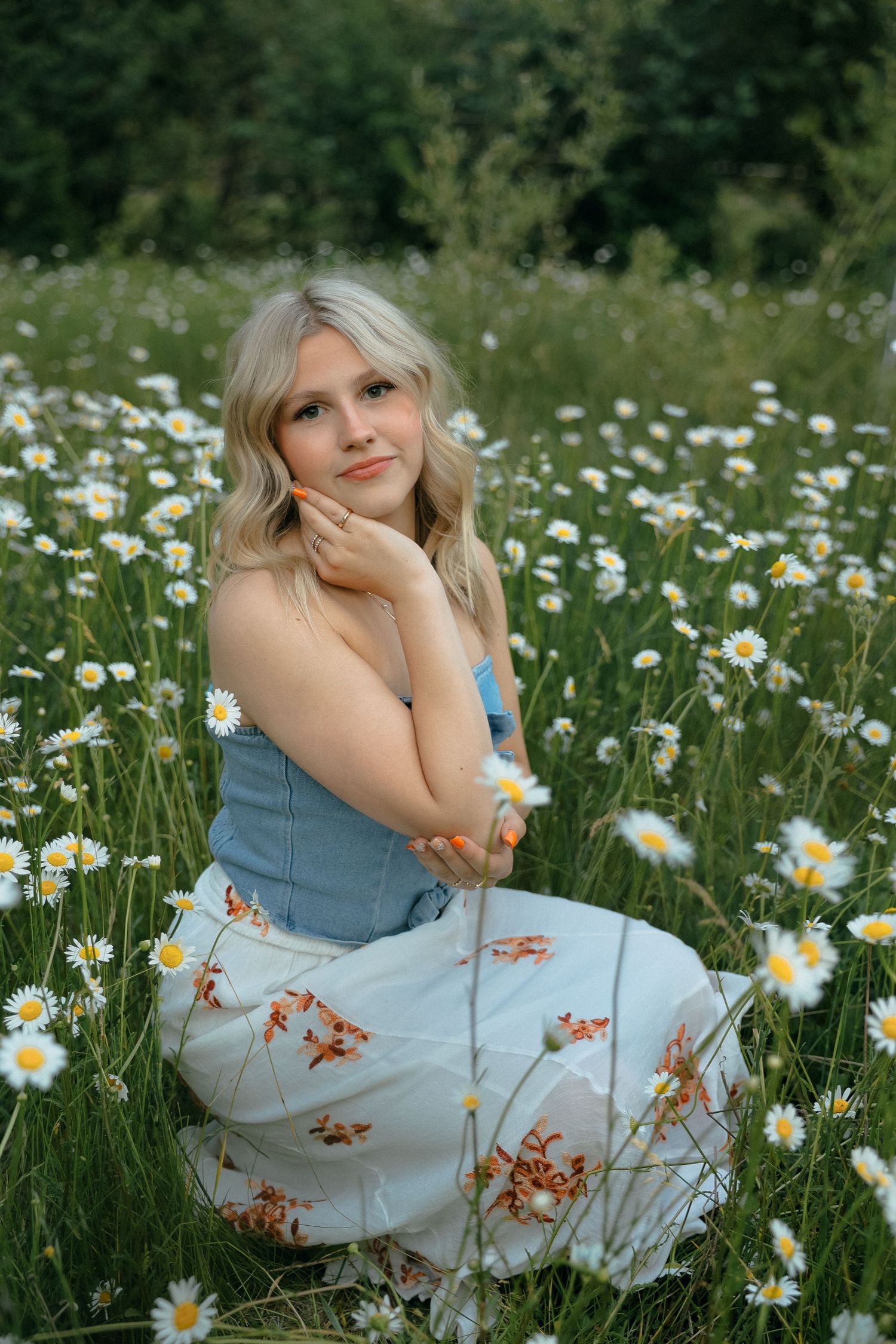 A person sits in a field of blooming daisies wearing a blue top and floral skirt on a summer day.