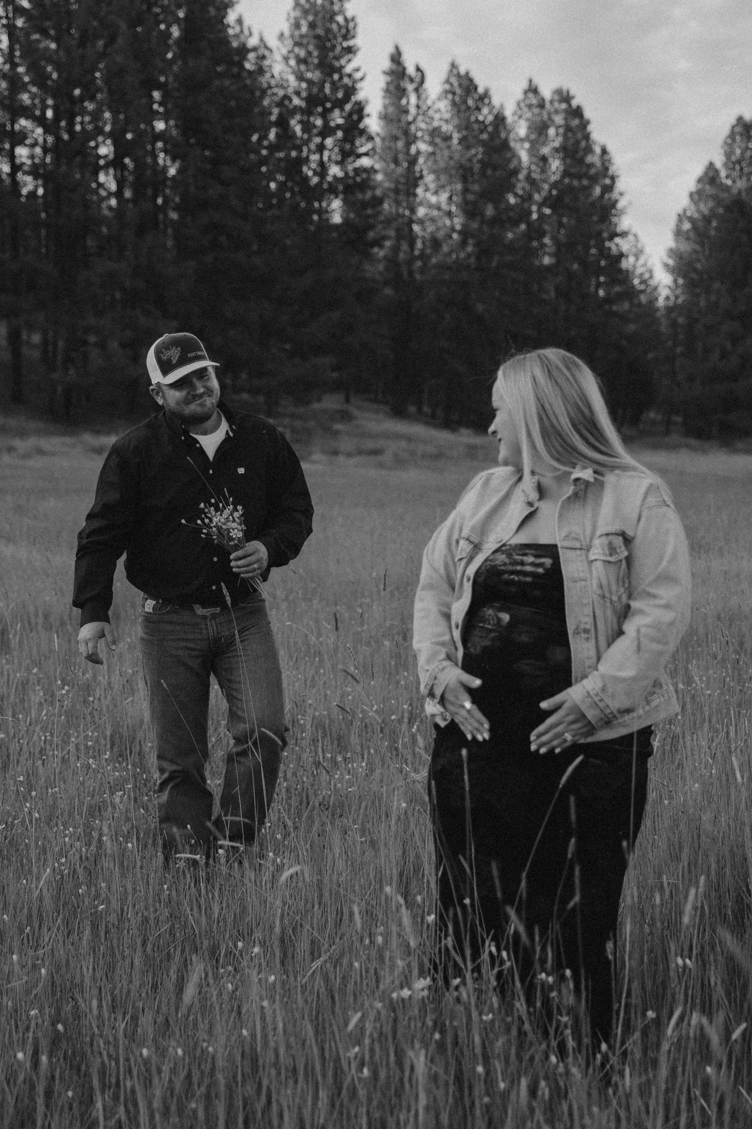 Black and white photo series showing a couple in casual attire standing together in a grassy meadow with pine trees in background.