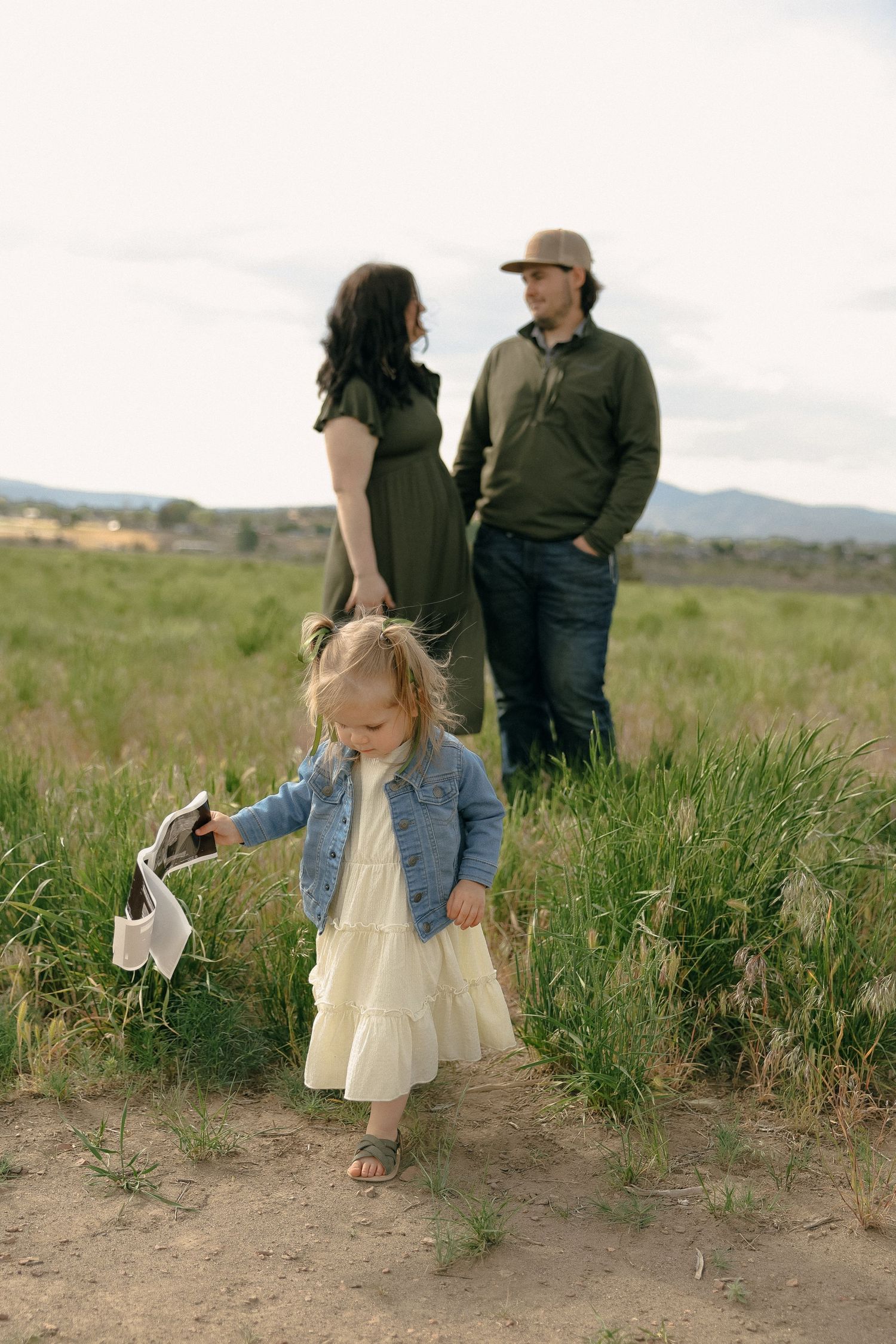 A small child in a denim jacket and dress explores a grassy field.