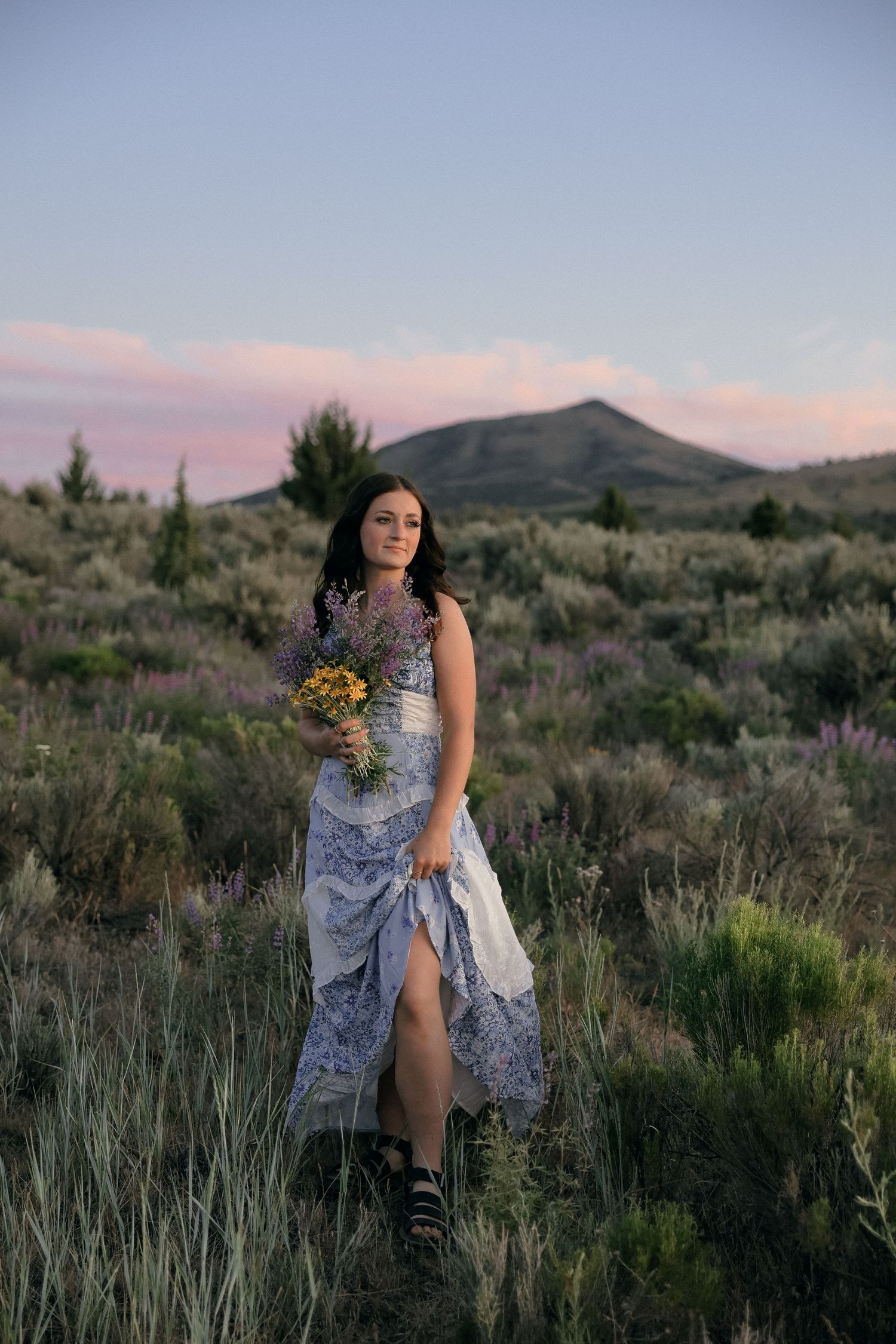 A person in a flowing blue dress holds wildflowers while standing in a desert landscape at dusk with mountains in background.