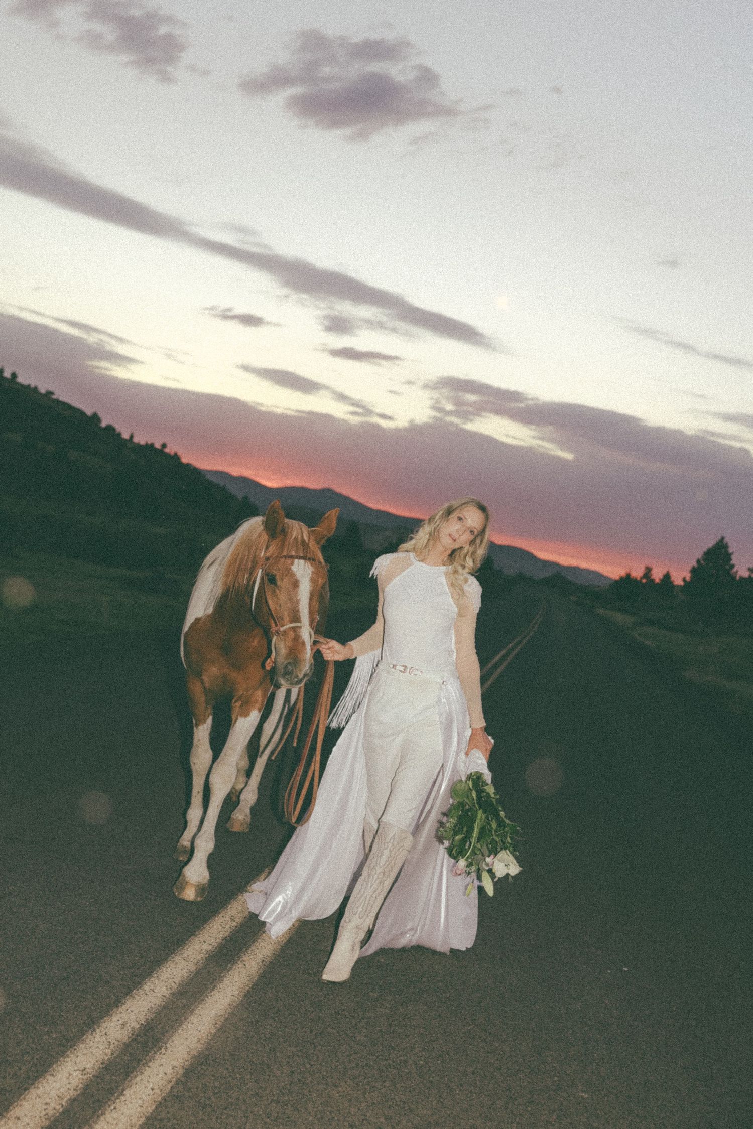 Person in wedding dress standing with horse during sunset in a scenic field.