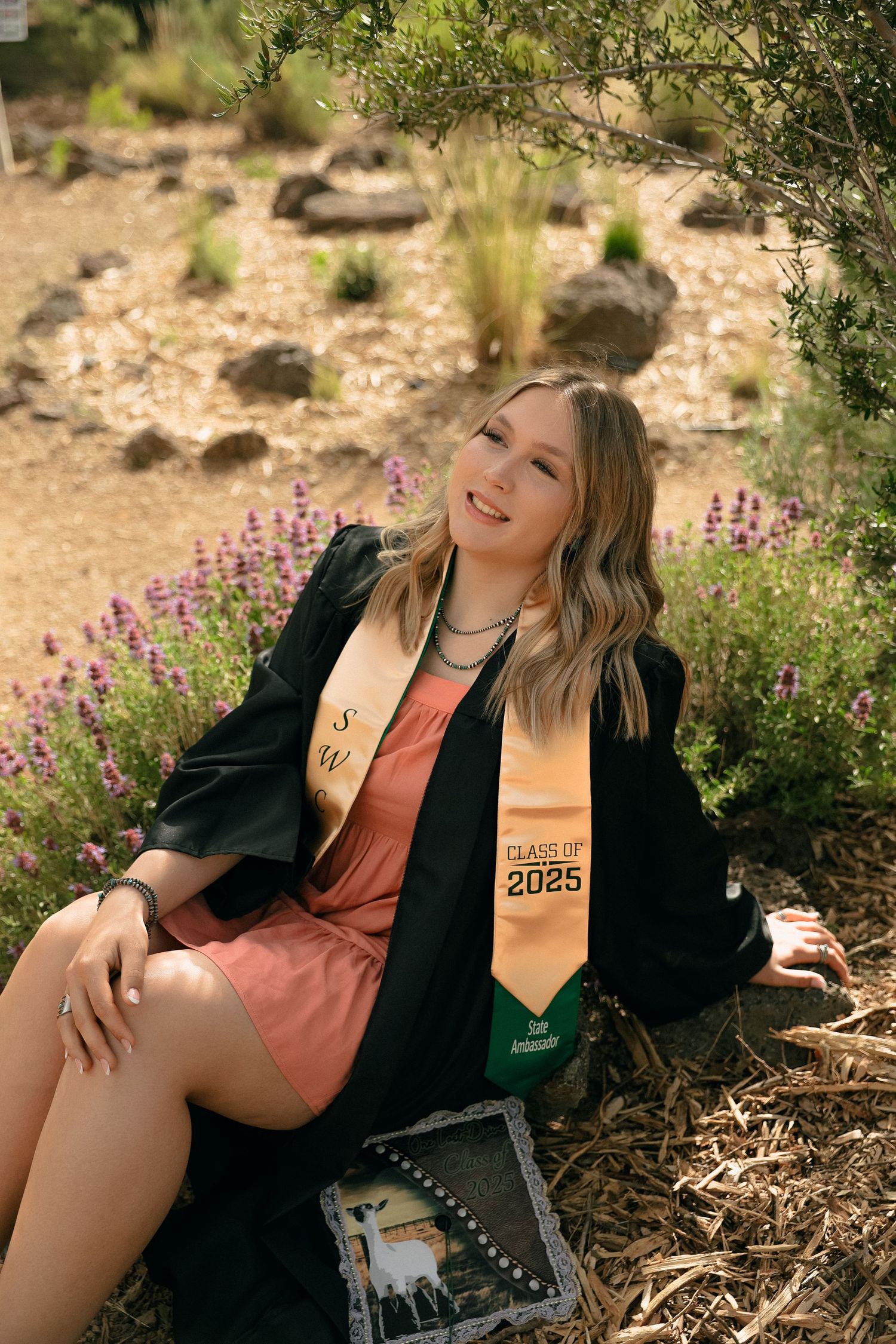 A graduate poses outdoors surrounded by desert landscaping and purple wildflowers.