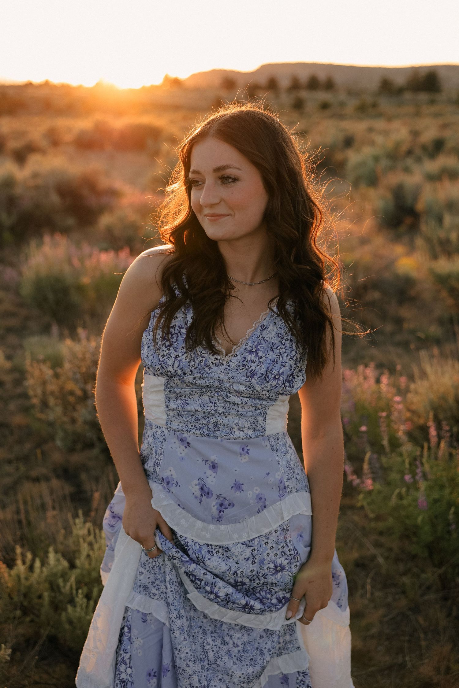 Person in a floral dress stands among desert brush during golden hour sunset in natural landscape.