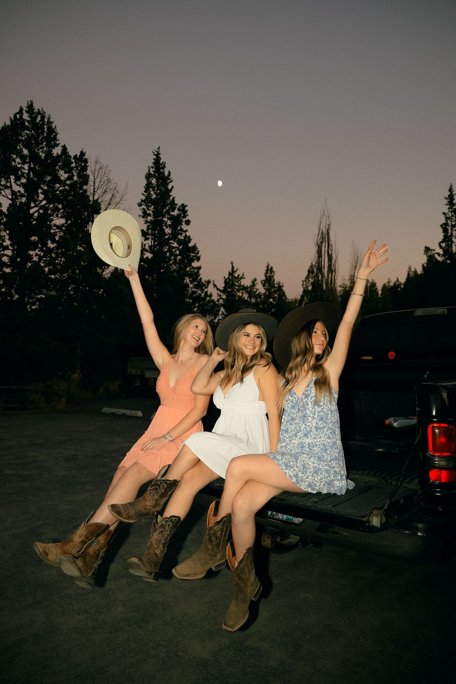 Silhouetted friends sitting on truck tailgate at dusk raising hats and arms in celebration.