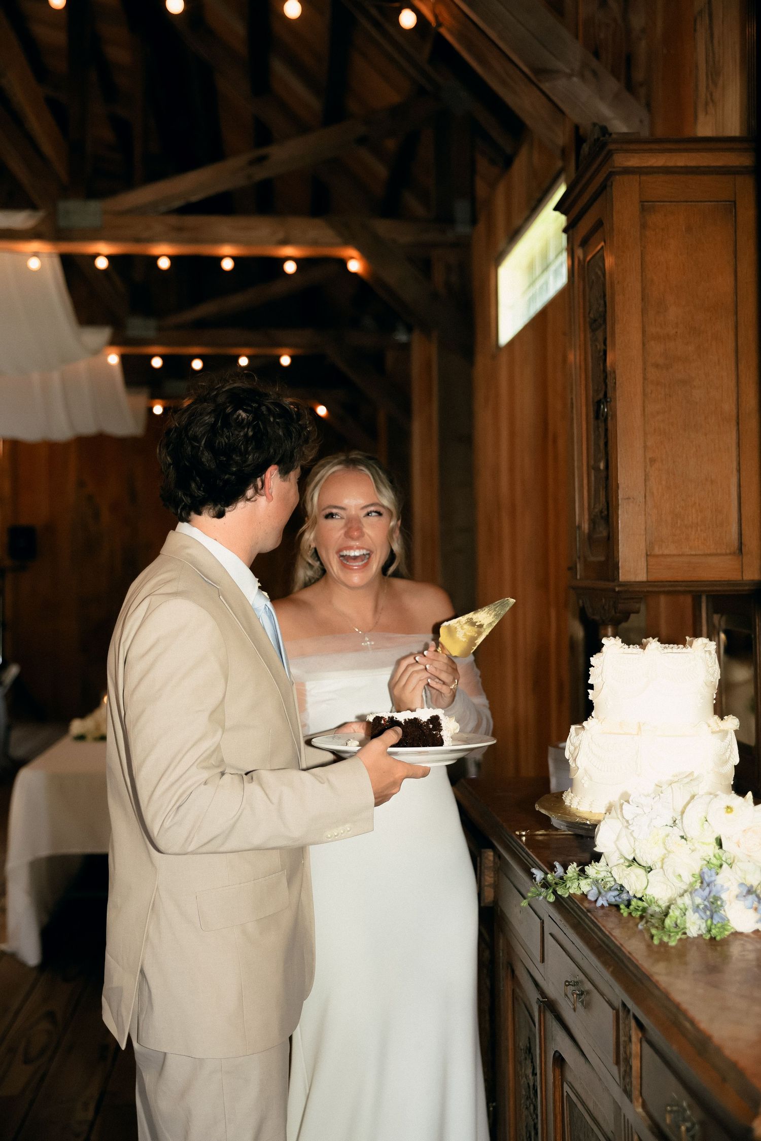 A couple shares a wedding cake at their rustic reception venue decorated with string lights.