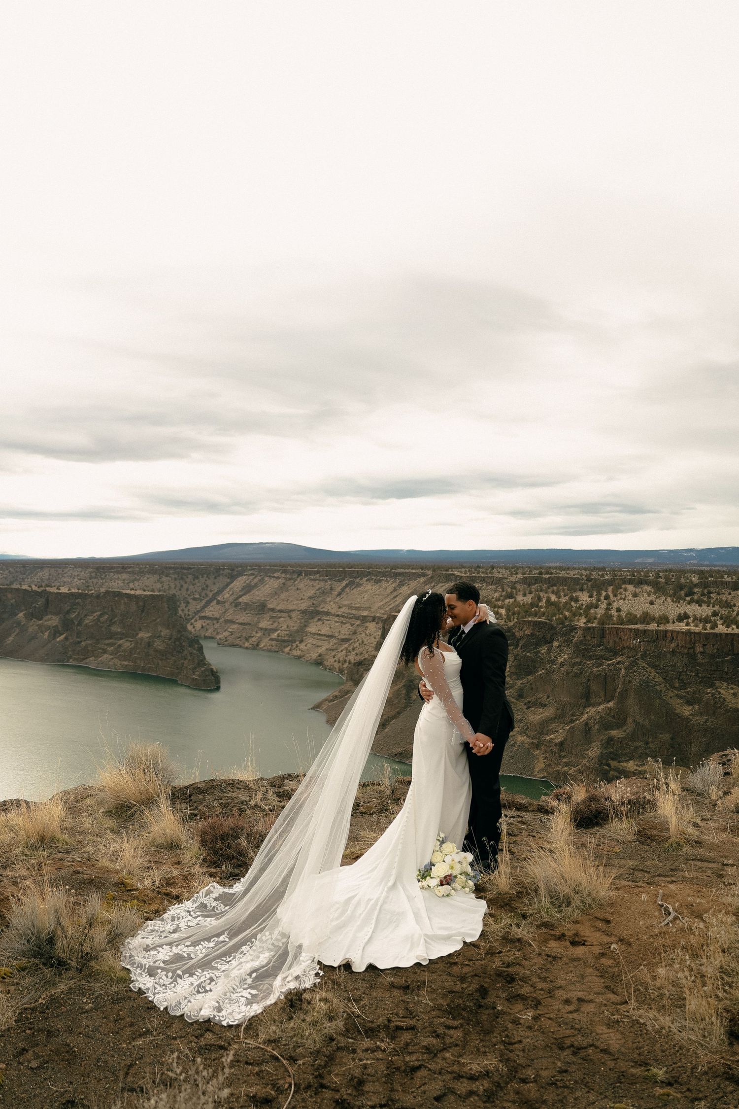Bride and groom embracing on desert cliffside with flowing veil.