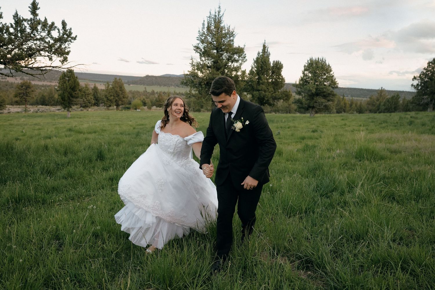 Newlyweds walking hand in hand through a lush green meadow with mountains in background.