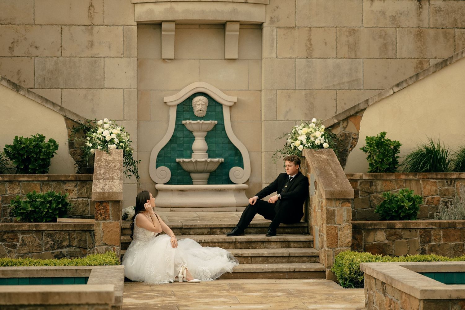 Wedding couple relaxing on ornate stone steps decorated with floral arrangements.