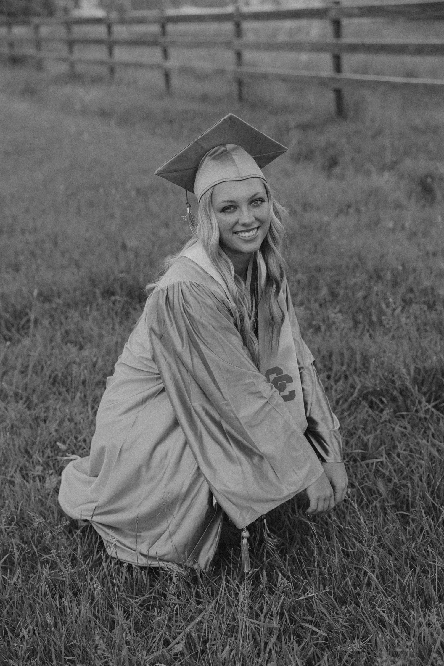 Graduate in cap and gown poses in field with wooden fence in black and white photo.
